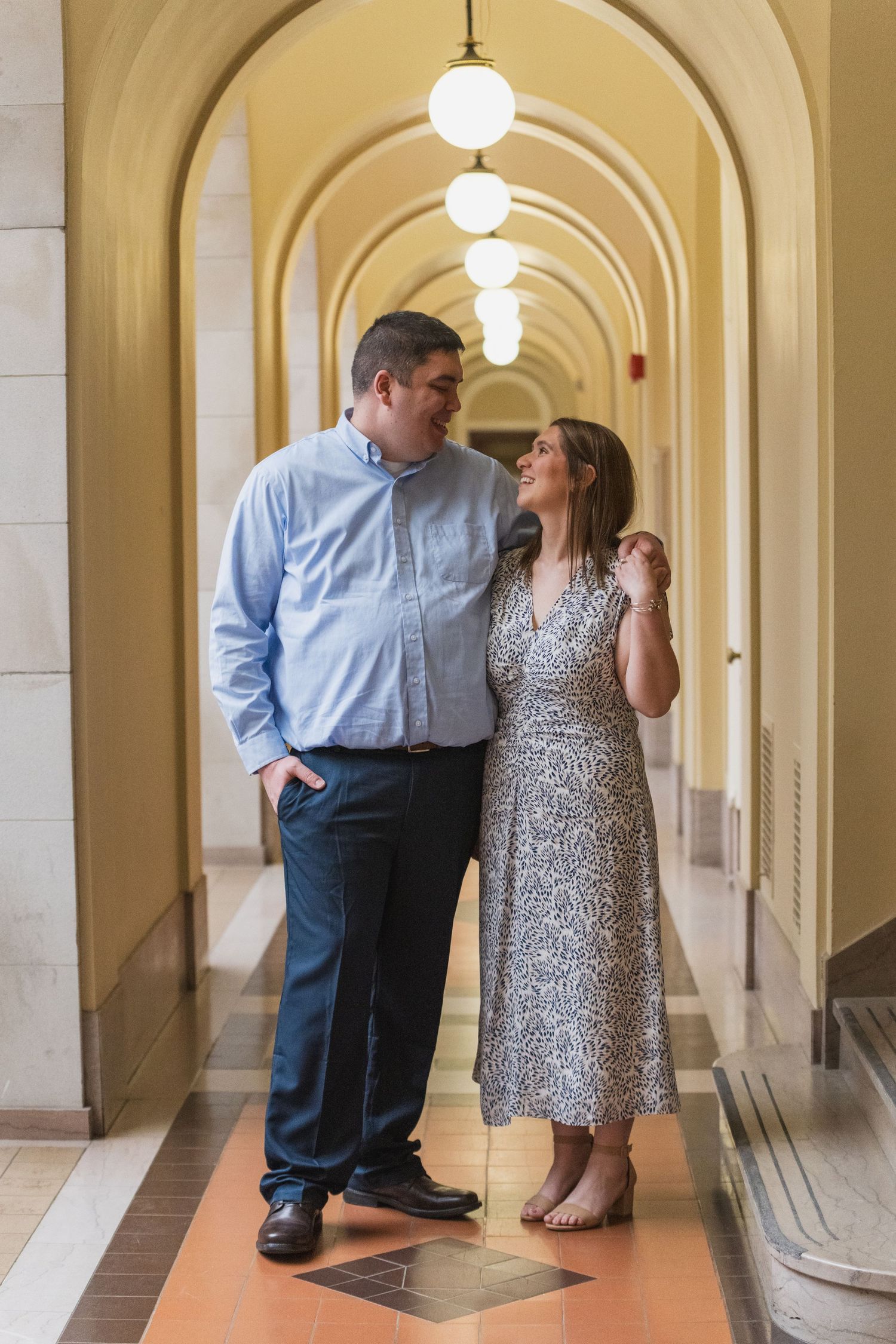A couple shares a tender moment in an elegant hallway with arched ceilings and columns.