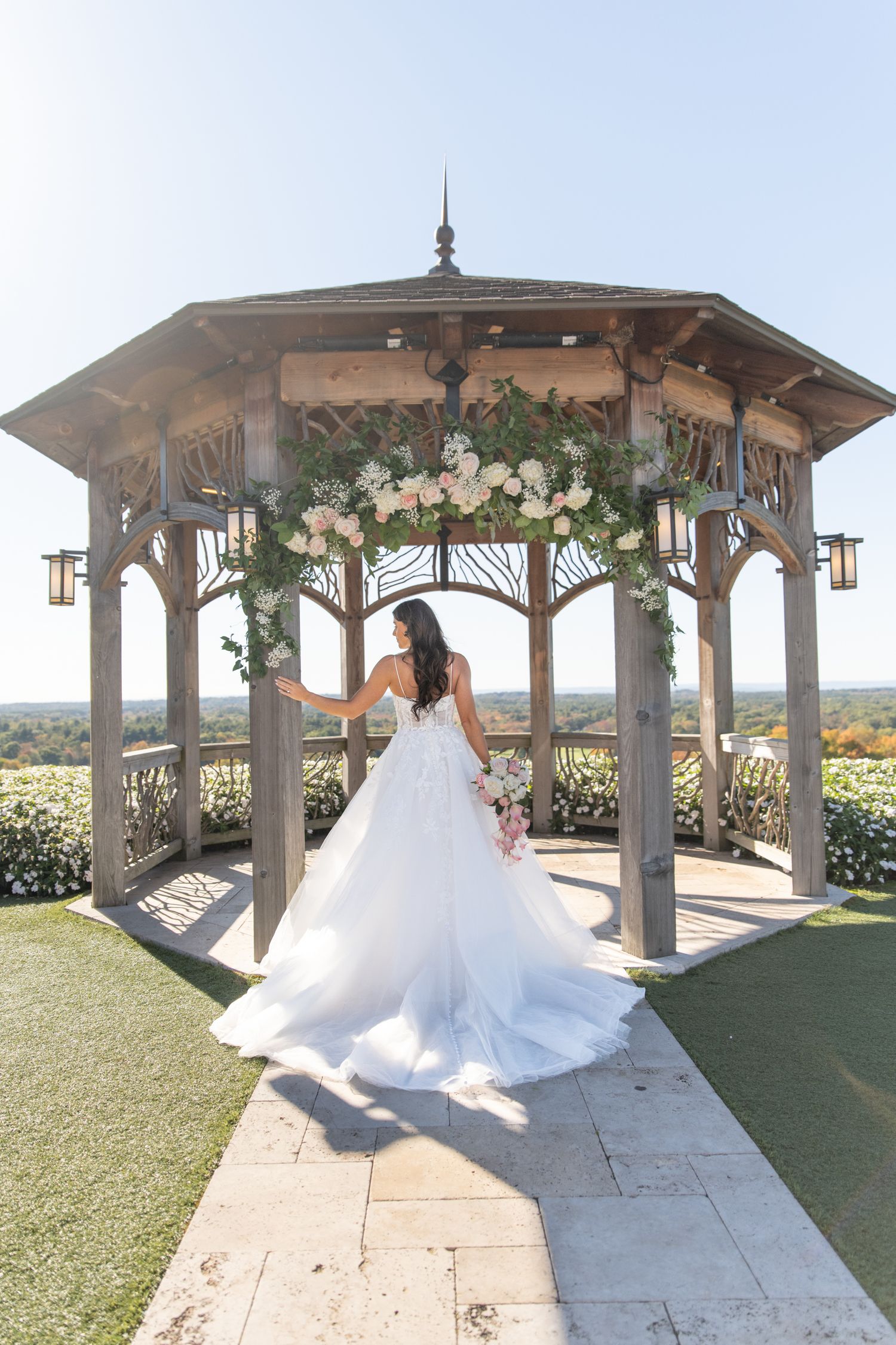 A bride in a flowing white wedding dress poses in a wooden gazebo overlooking a scenic landscape at sunset.