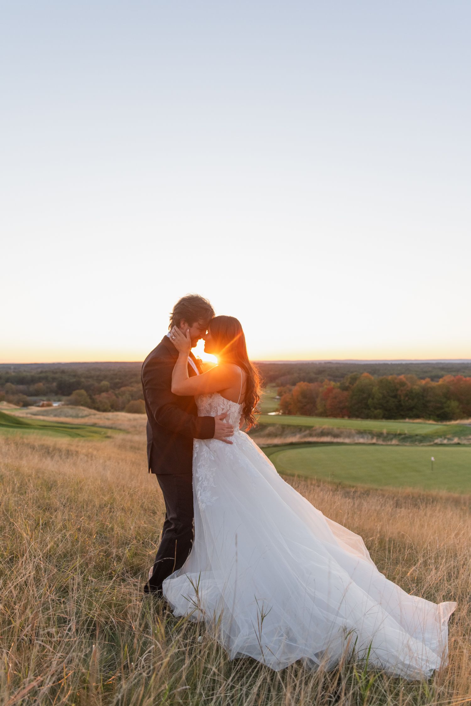 A couple in wedding attire share a romantic kiss at sunset on a hilltop overlooking scenic rolling meadows.