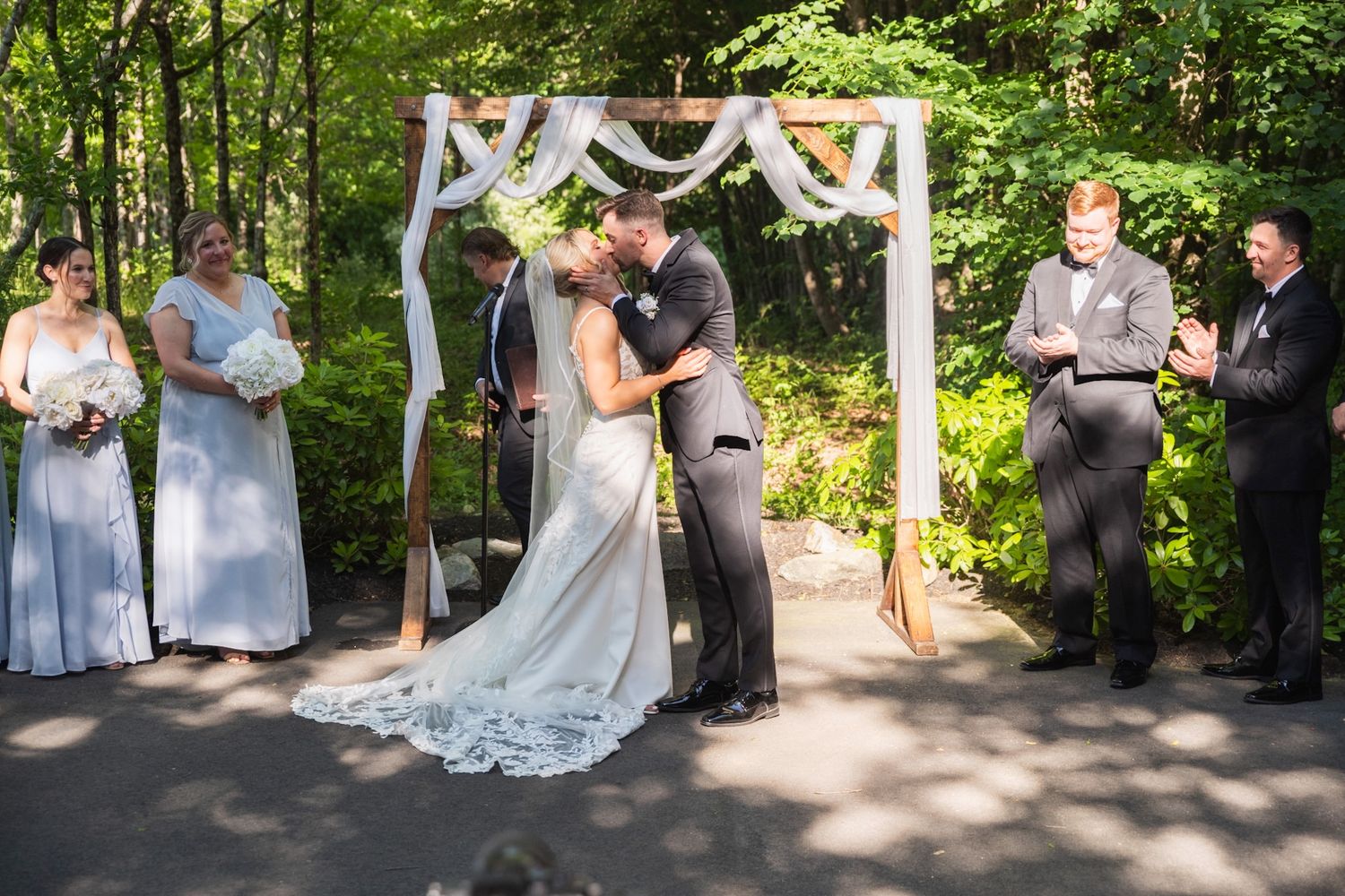 Outdoor wedding ceremony under white draped arch with wedding party in soft gray attire.