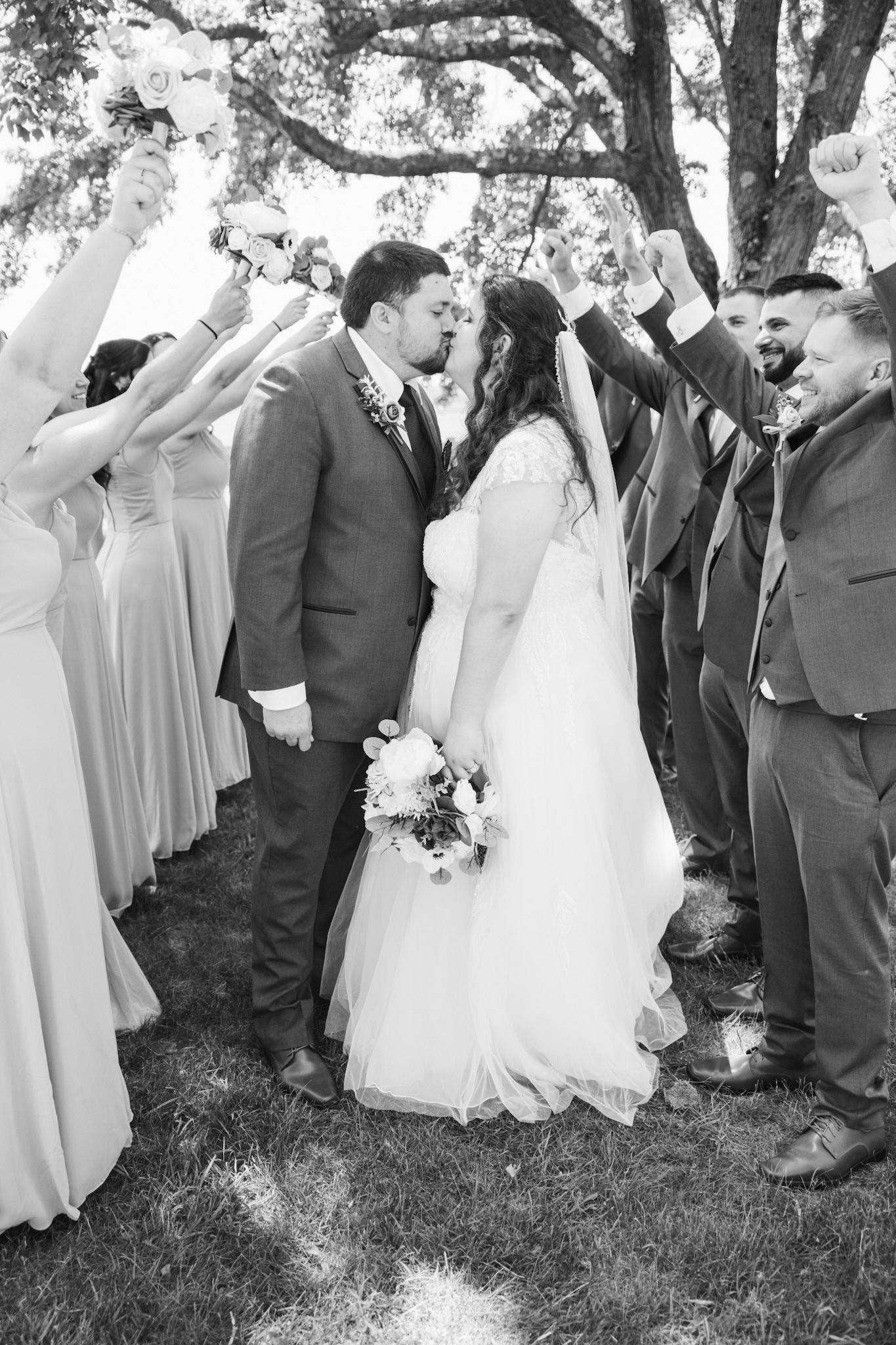 Wedding party celebrating with joyful reactions during outdoor group photo session.