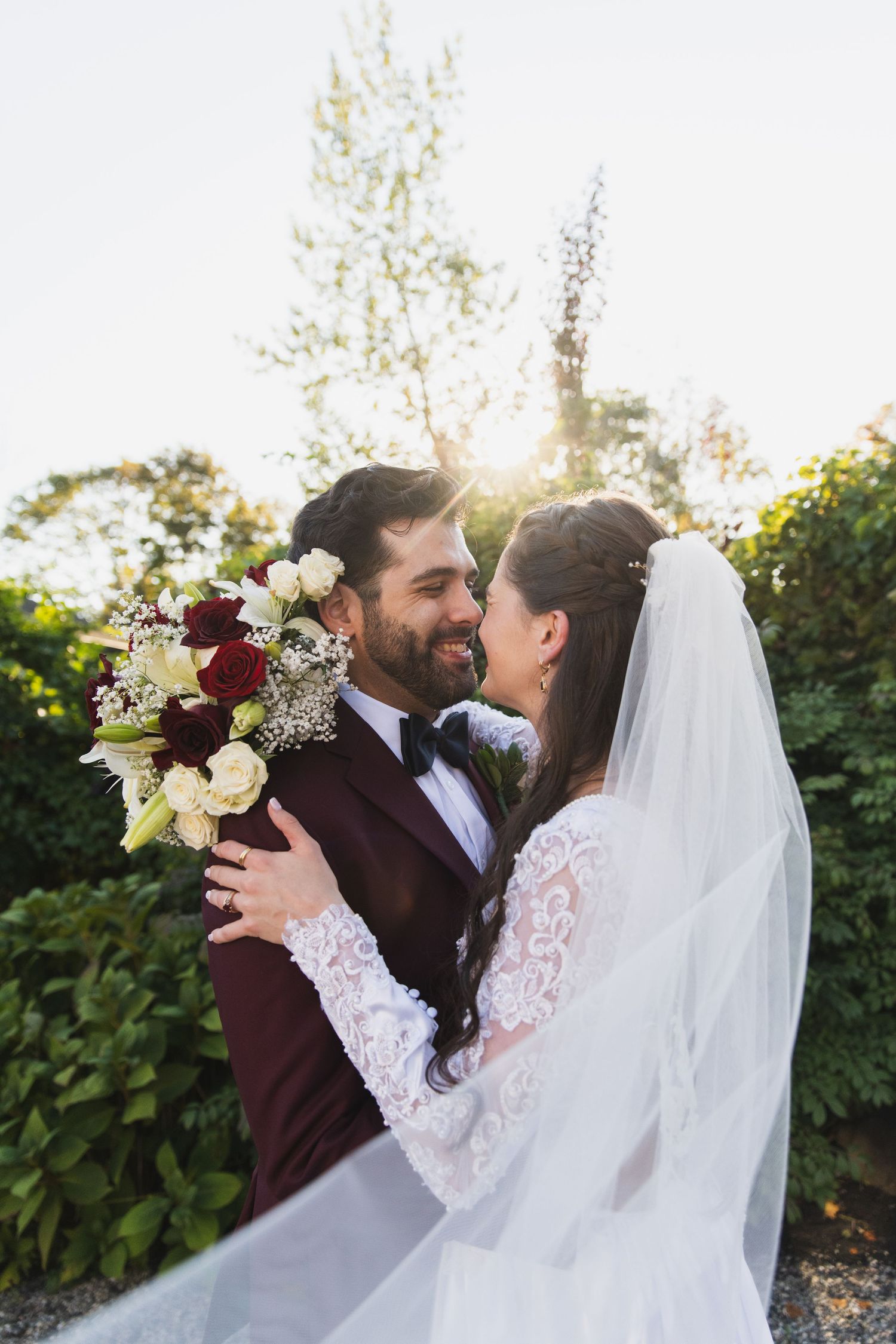A romantic wedding couple embraces outdoors in a garden setting with sunlight filtering through trees.