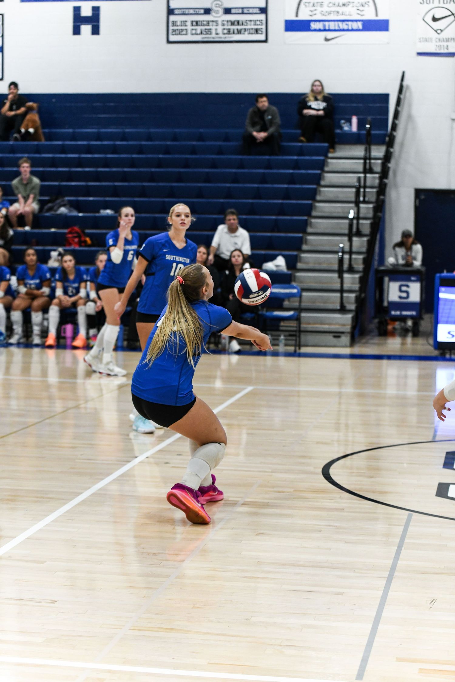 Female volleyball player in blue uniform performs a dig during an indoor high school volleyball game with teammates and spectators.
