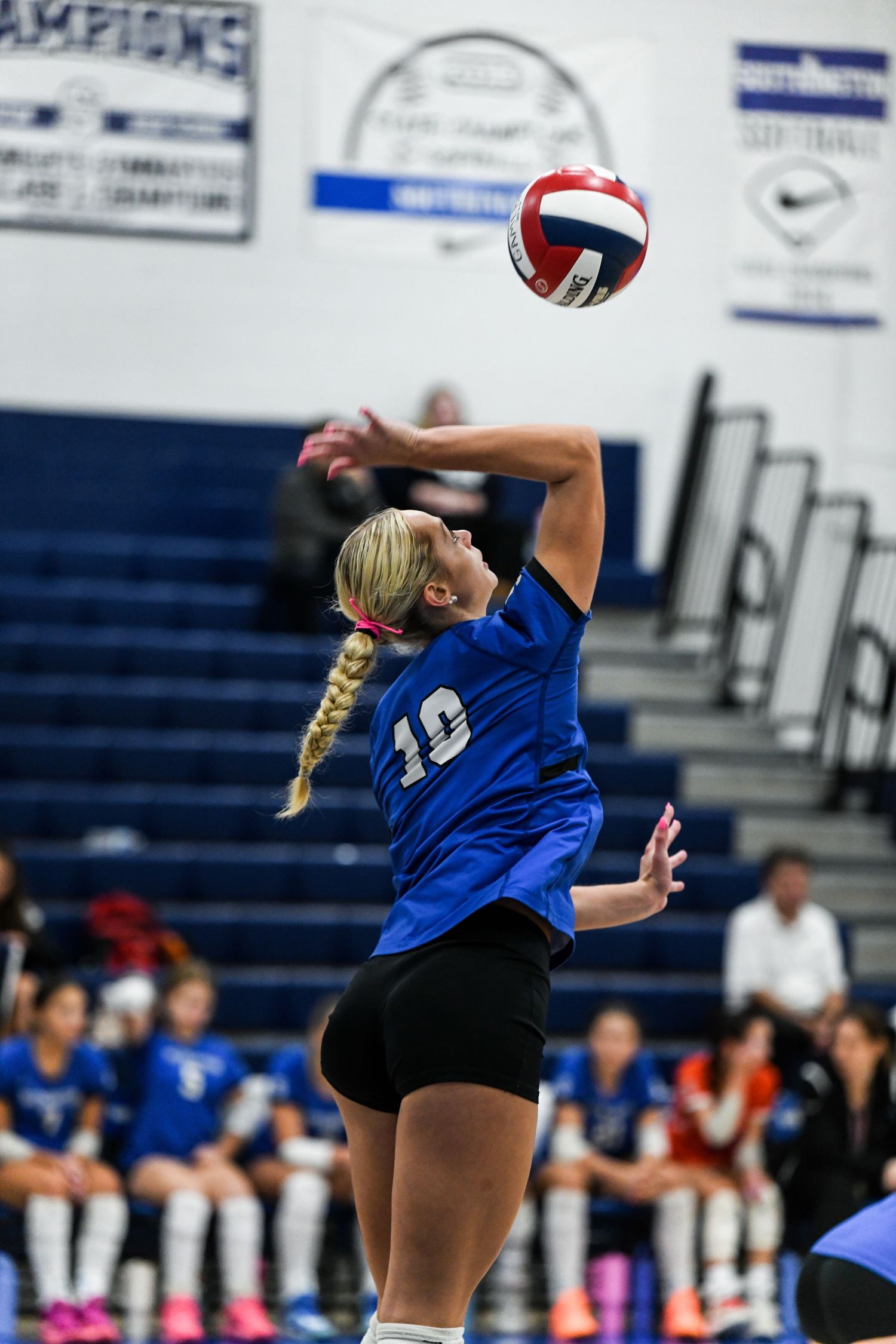 Female volleyball player in blue jersey #10 jumping to serve during an indoor high school volleyball game.