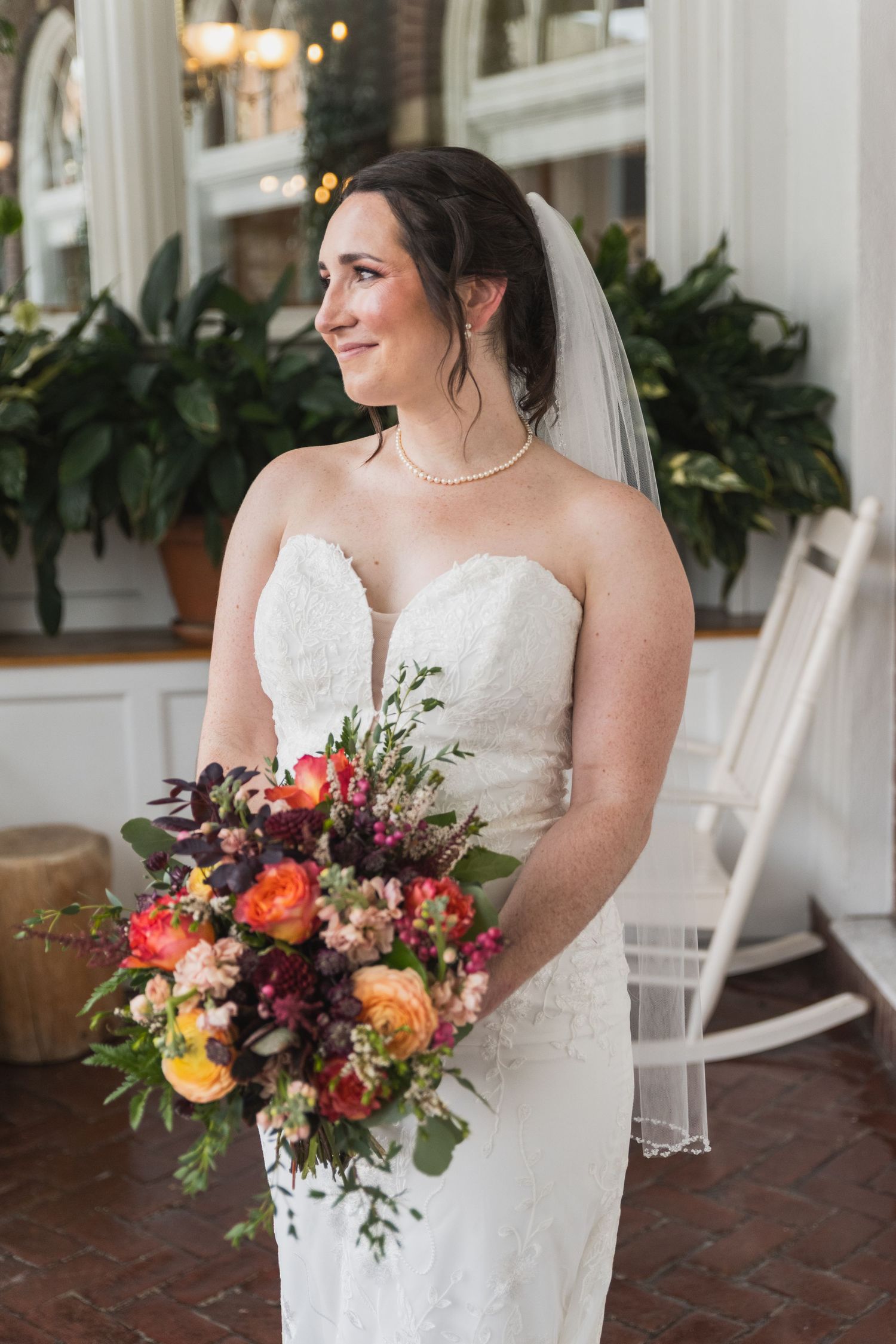A bride in a white lace gown holds a colorful autumn bouquet while sitting on a white rocking chair on a brick porch.