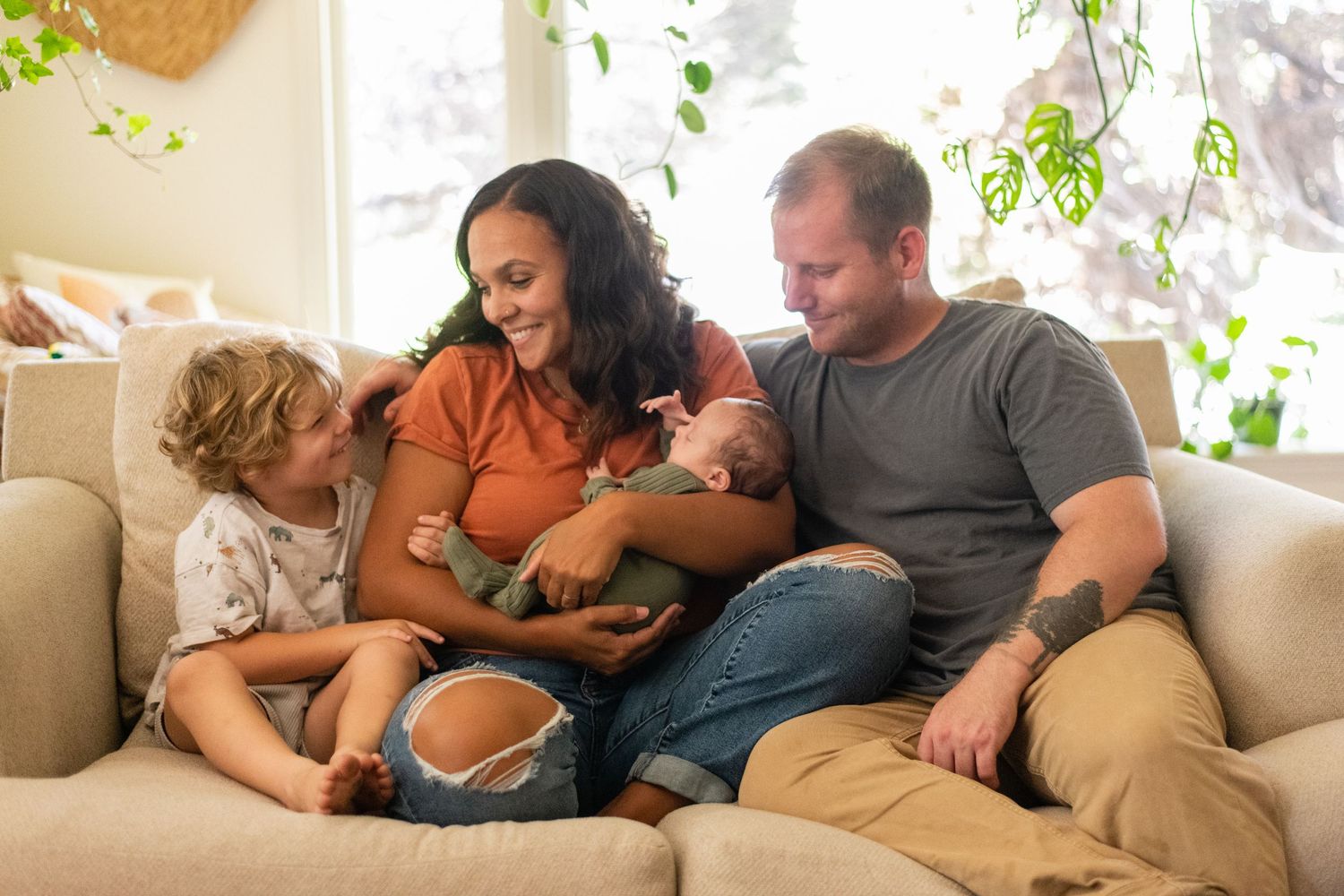 Intimate family moment captured on a cozy white couch with hanging plants in background.