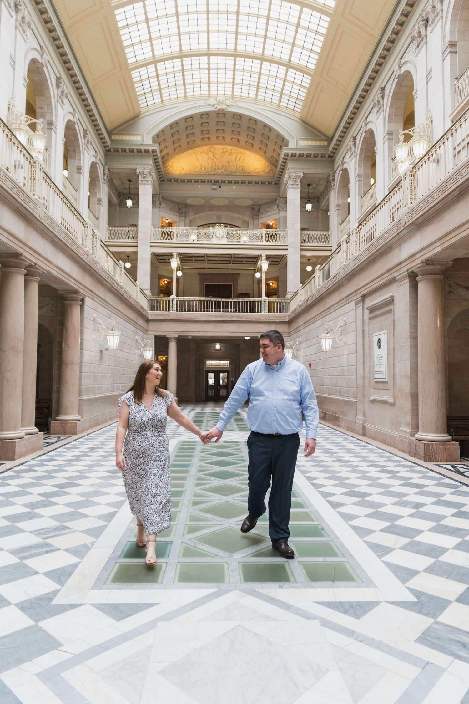 A couple walks hand in hand through a grand historic building with ornate architecture and checkered floors.