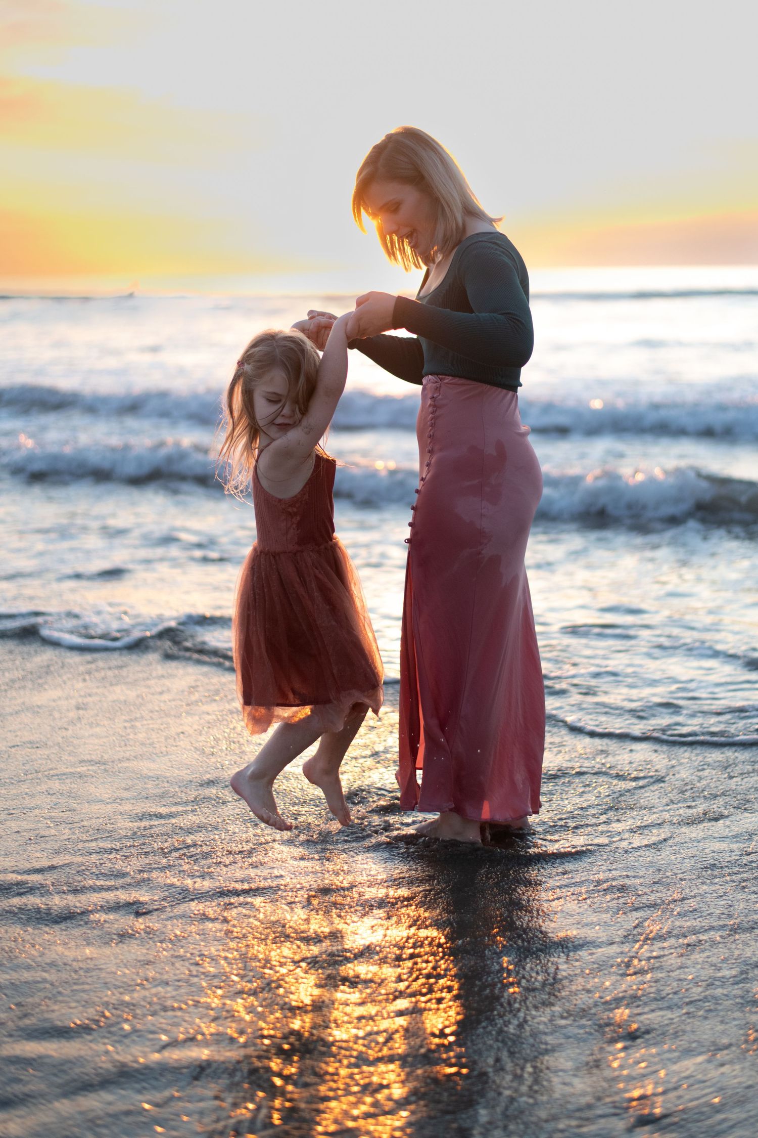 Two figures dance joyfully in the ocean waves during golden hour, creating rippling reflections.