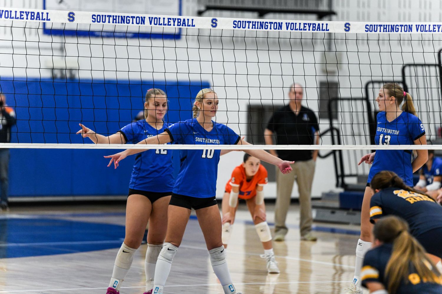 Two Southington volleyball players in blue uniforms stand ready at the net during a competitive indoor volleyball match.