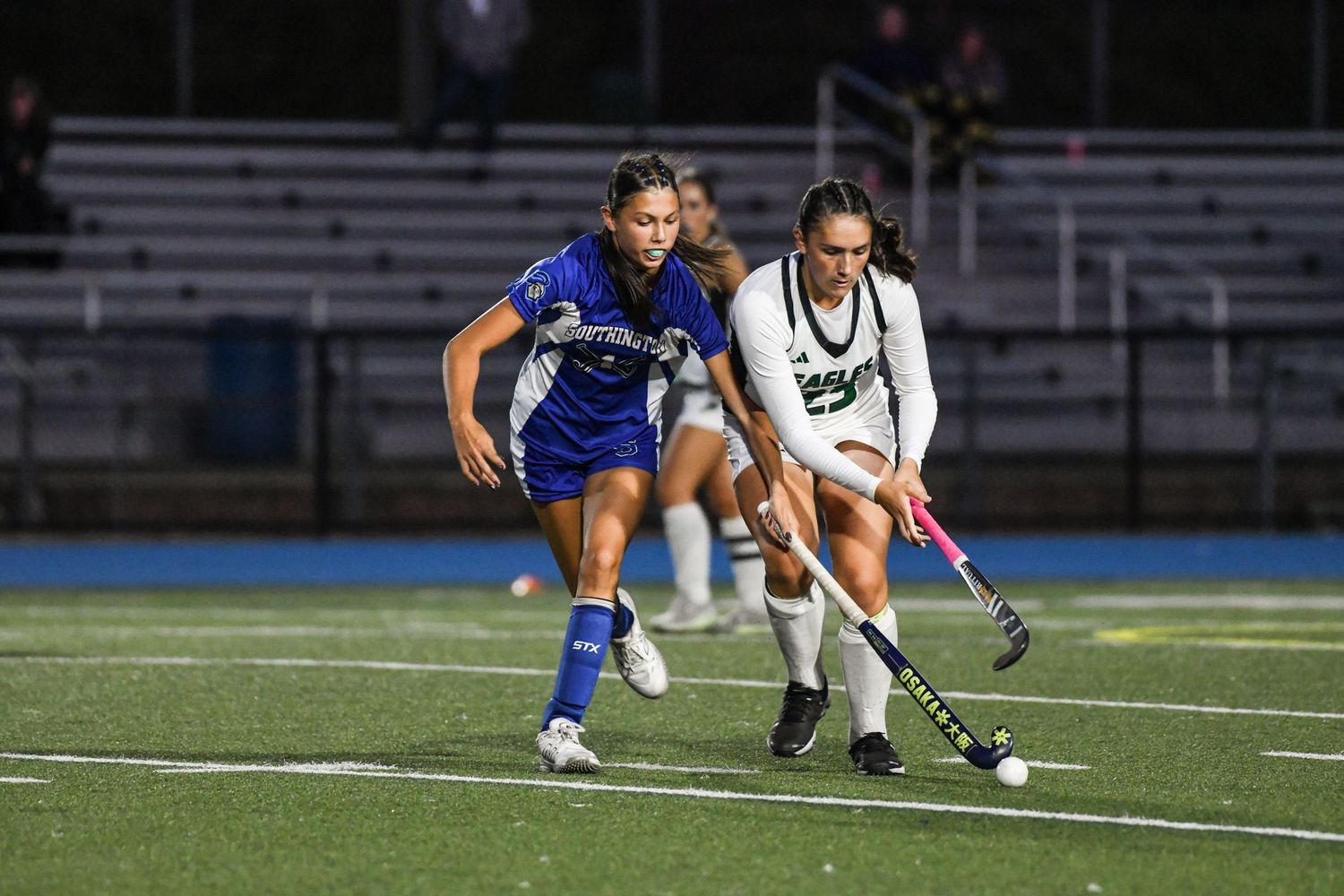 Two female field hockey players compete on a lit turf field at night, one in blue and one in white uniforms.