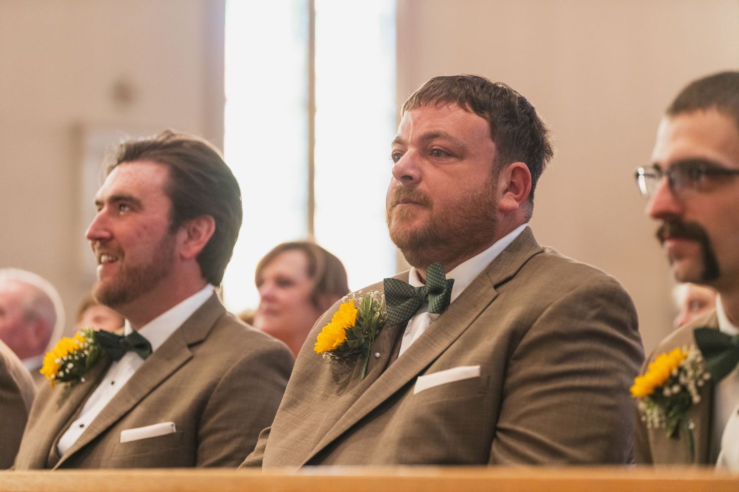 Groomsmen in brown suits with yellow boutonnieres seated during ceremony.