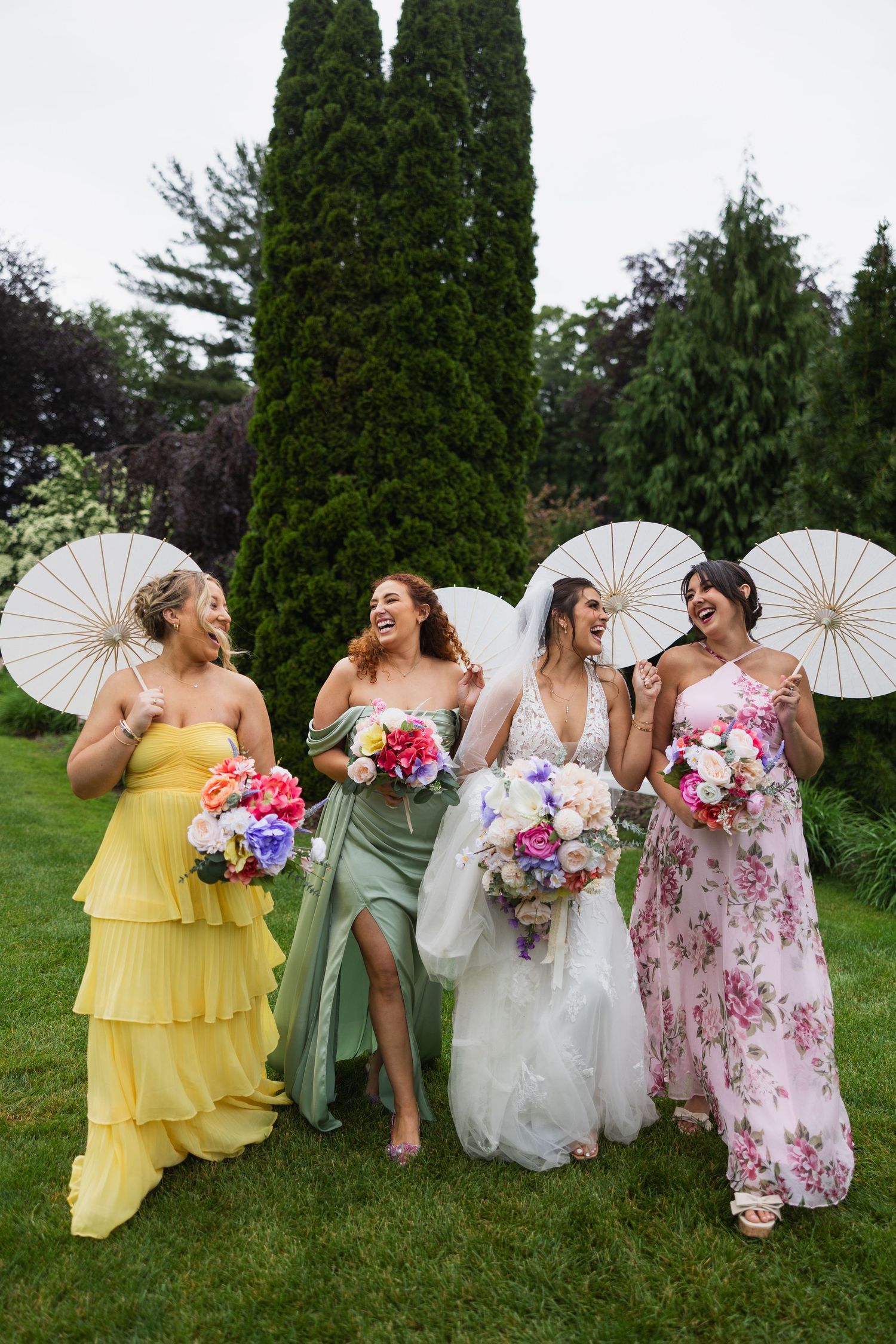 Wedding party poses with white parasols against green garden backdrop.