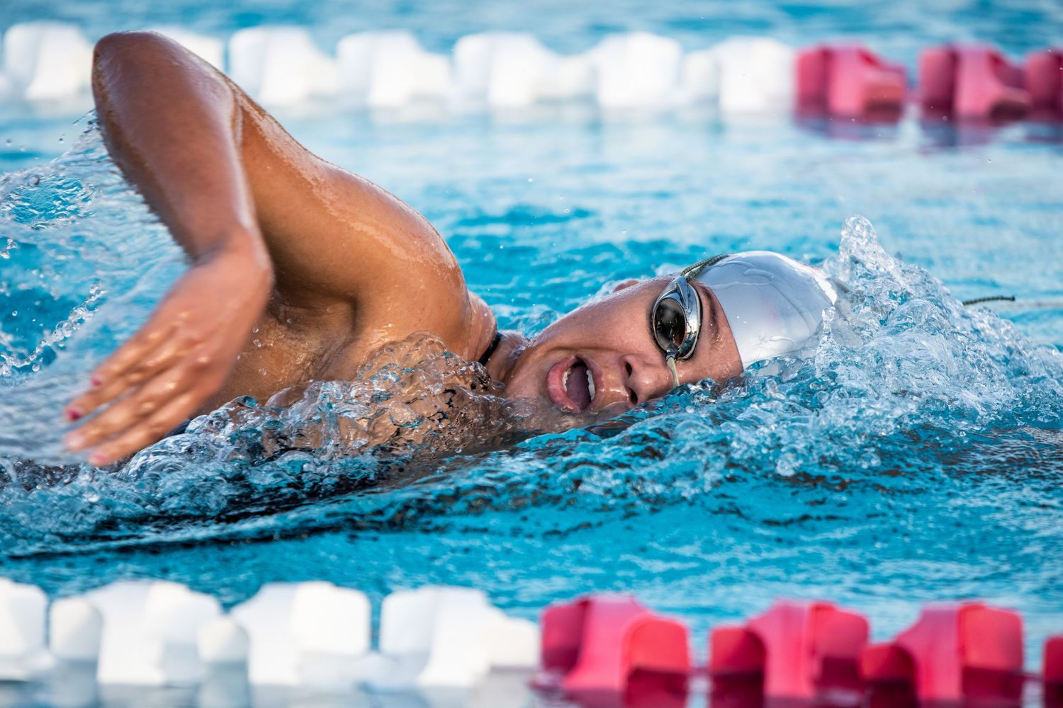Competitive swimmer doing freestyle stroke in a pool, wearing goggles and white swim cap, with splashing water.