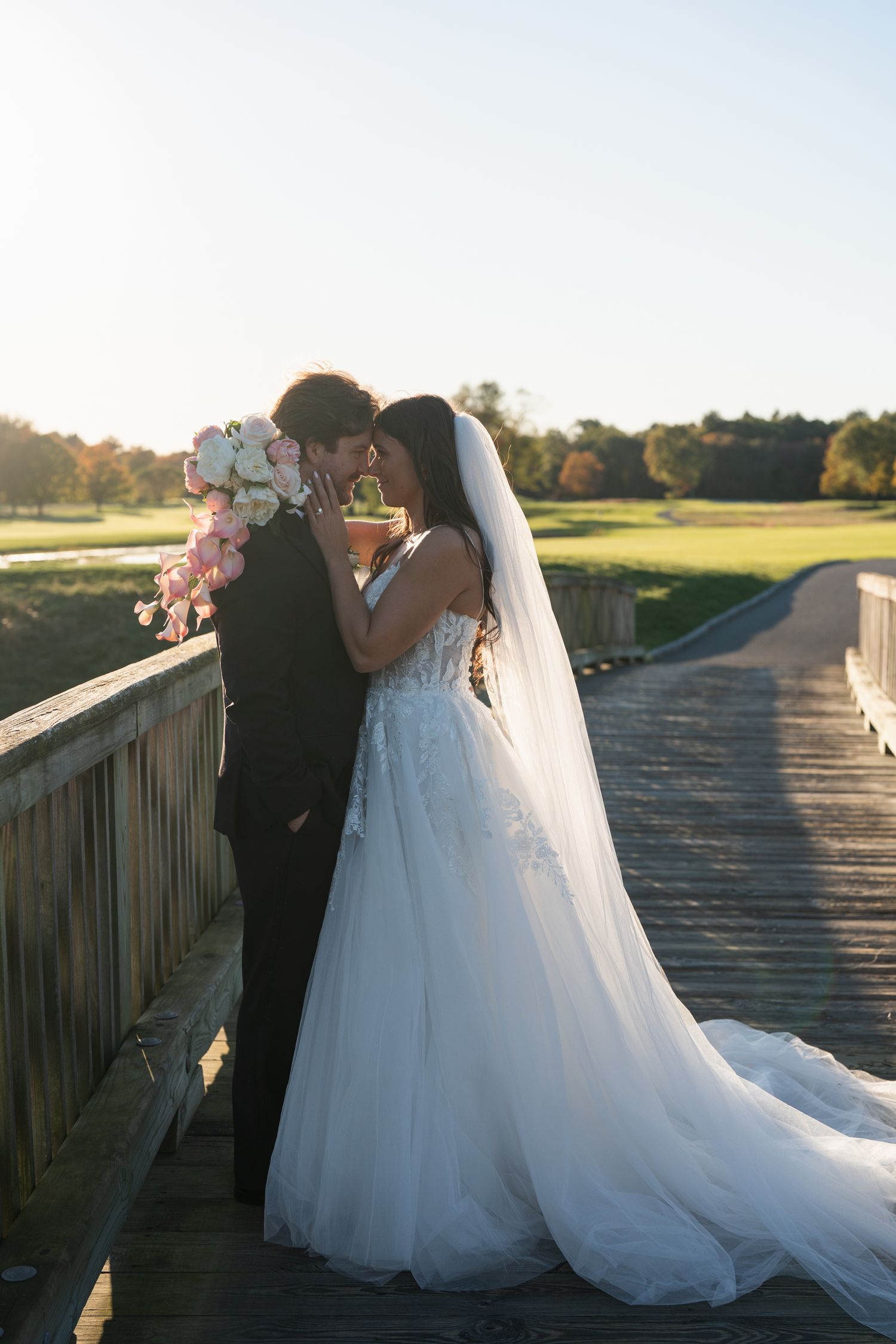 A romantic wedding couple embraces on a wooden bridge at sunset, with the bride wearing a flowing white gown and veil.