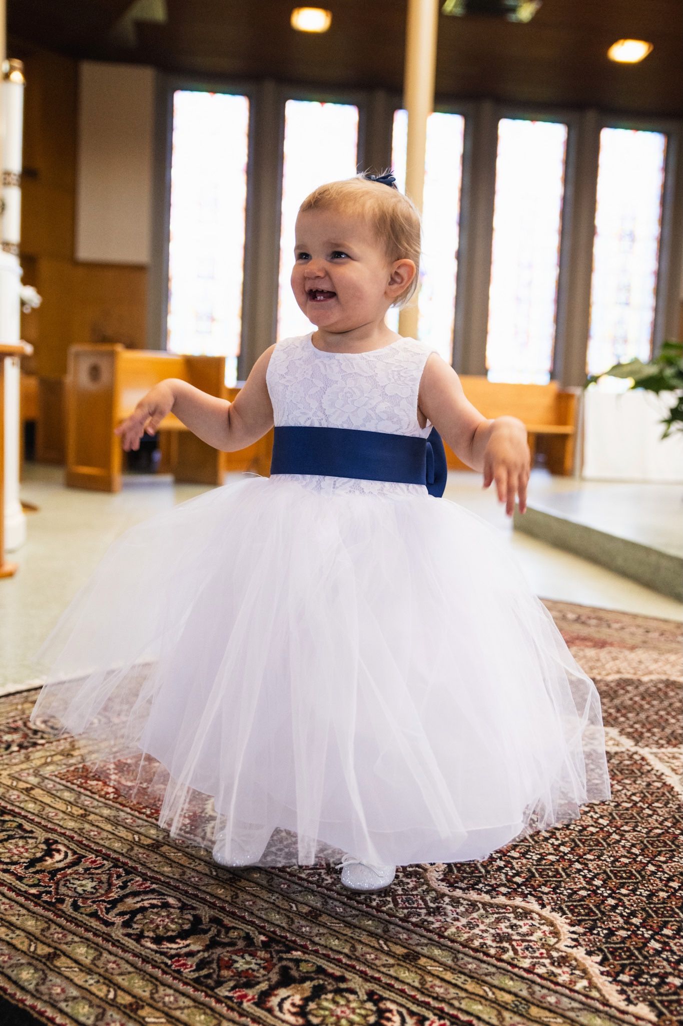Small child in white tulle dress crawls on ornate carpet during wedding celebration.