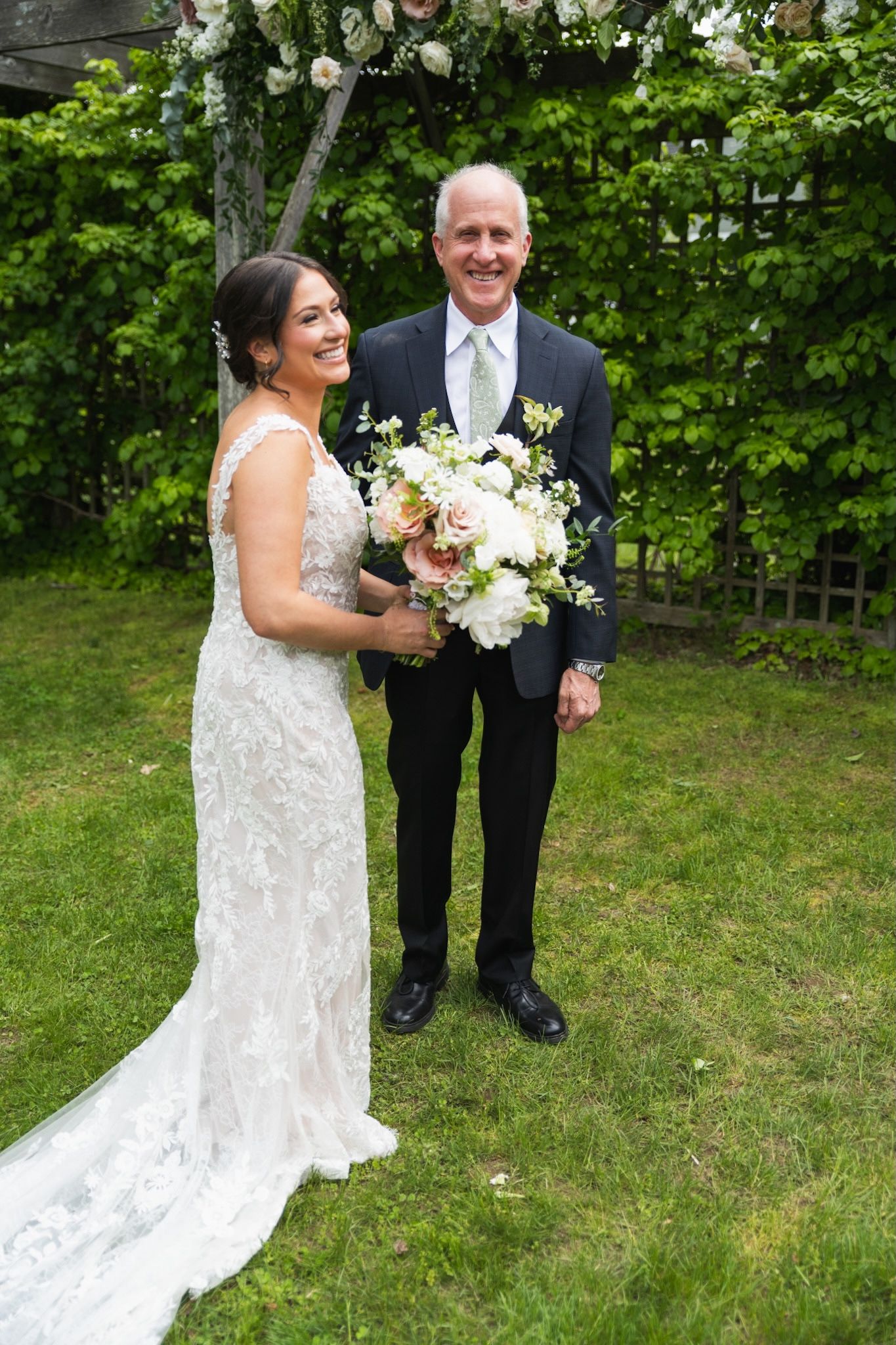 A bride in a white lace dress holding a bouquet poses on lush green grass for wedding photos.