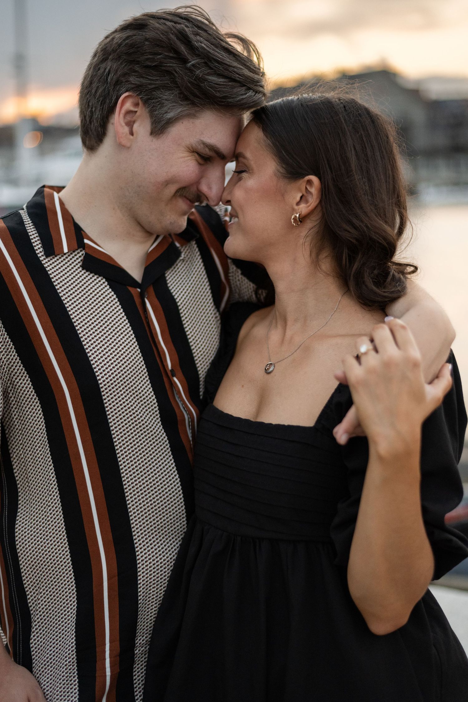 A couple shares intimate moments at sunset along a waterfront, wearing black and striped outfits.