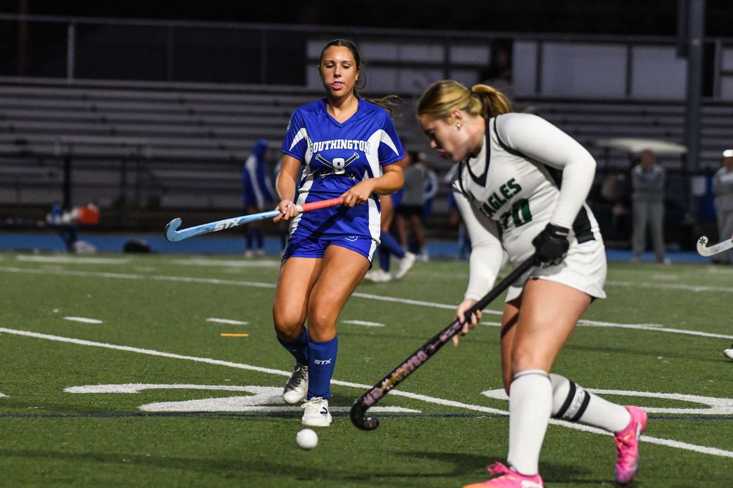 Two female field hockey players in blue and green uniforms compete for the ball on a turf field.