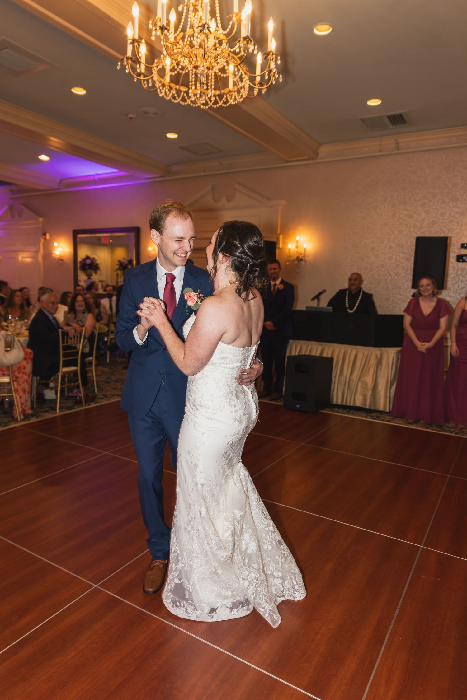 Newlyweds share their first dance on a wooden floor under elegant chandeliers at their wedding reception.