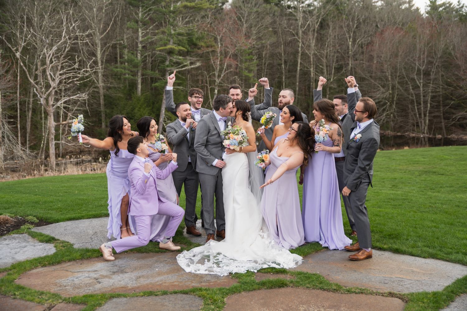 Wedding party poses playfully on stone patio with bridesmaids in lavender dresses.