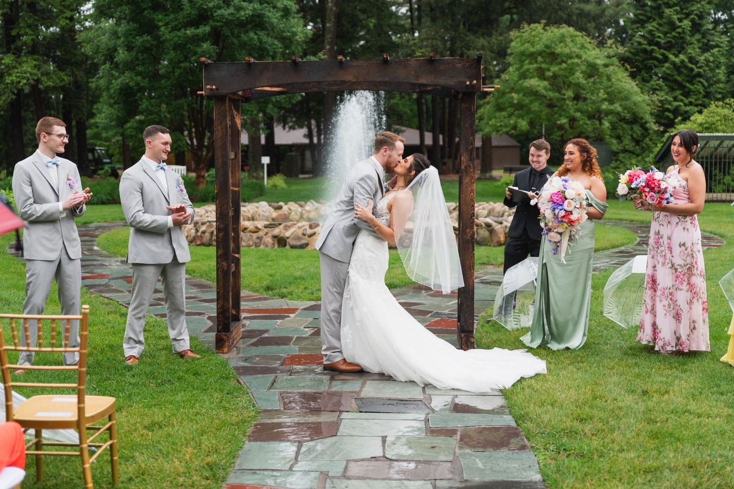 Wedding party celebrates on outdoor stone path surrounded by green gardens.