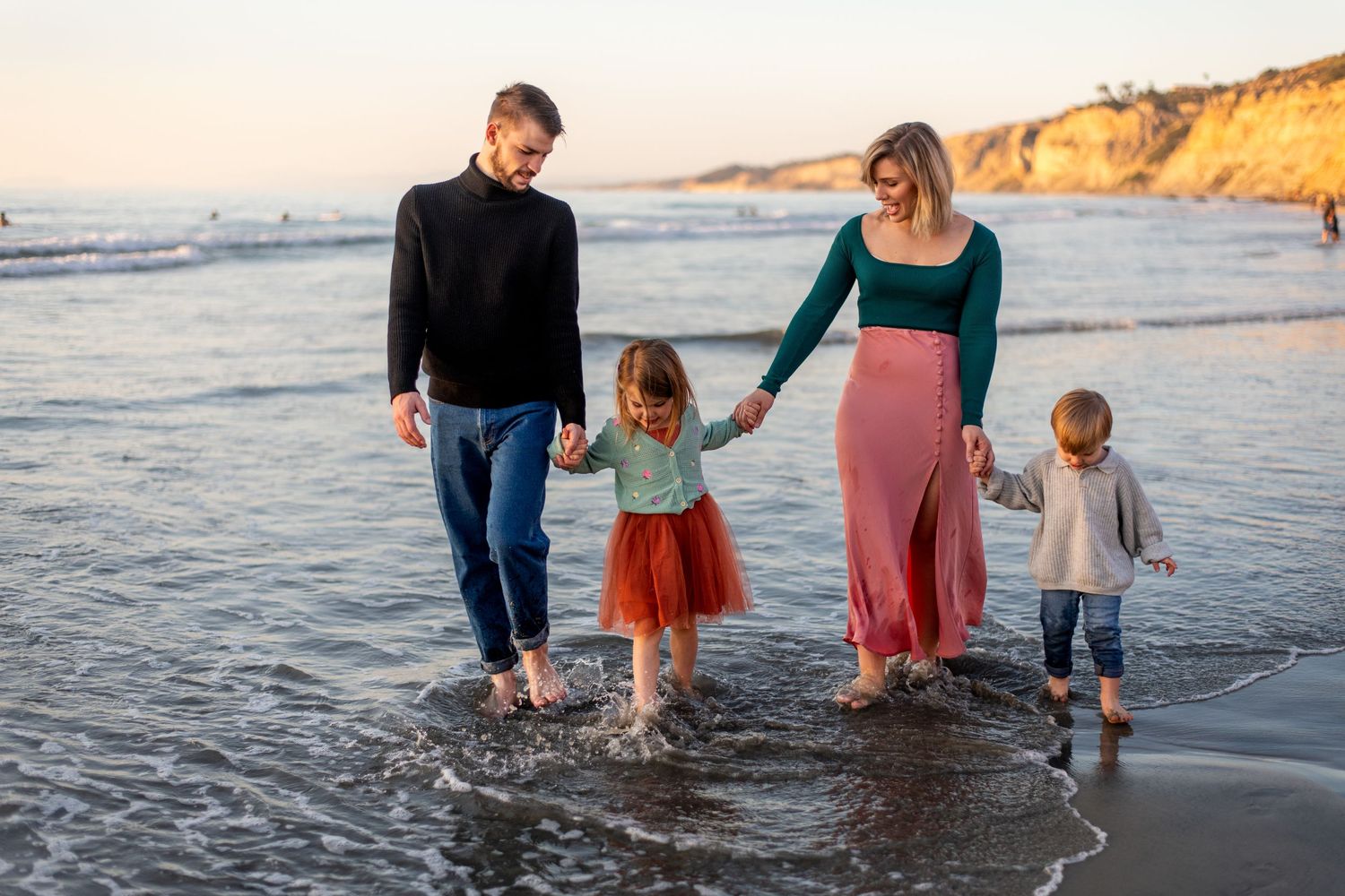 Family walking together along beach shoreline at sunset holding hands.