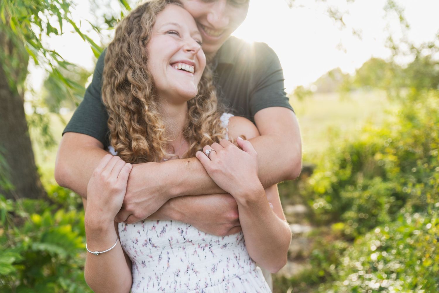 Happy couple embraces and laughs together in lush summer garden with sunlit greenery and flowers.