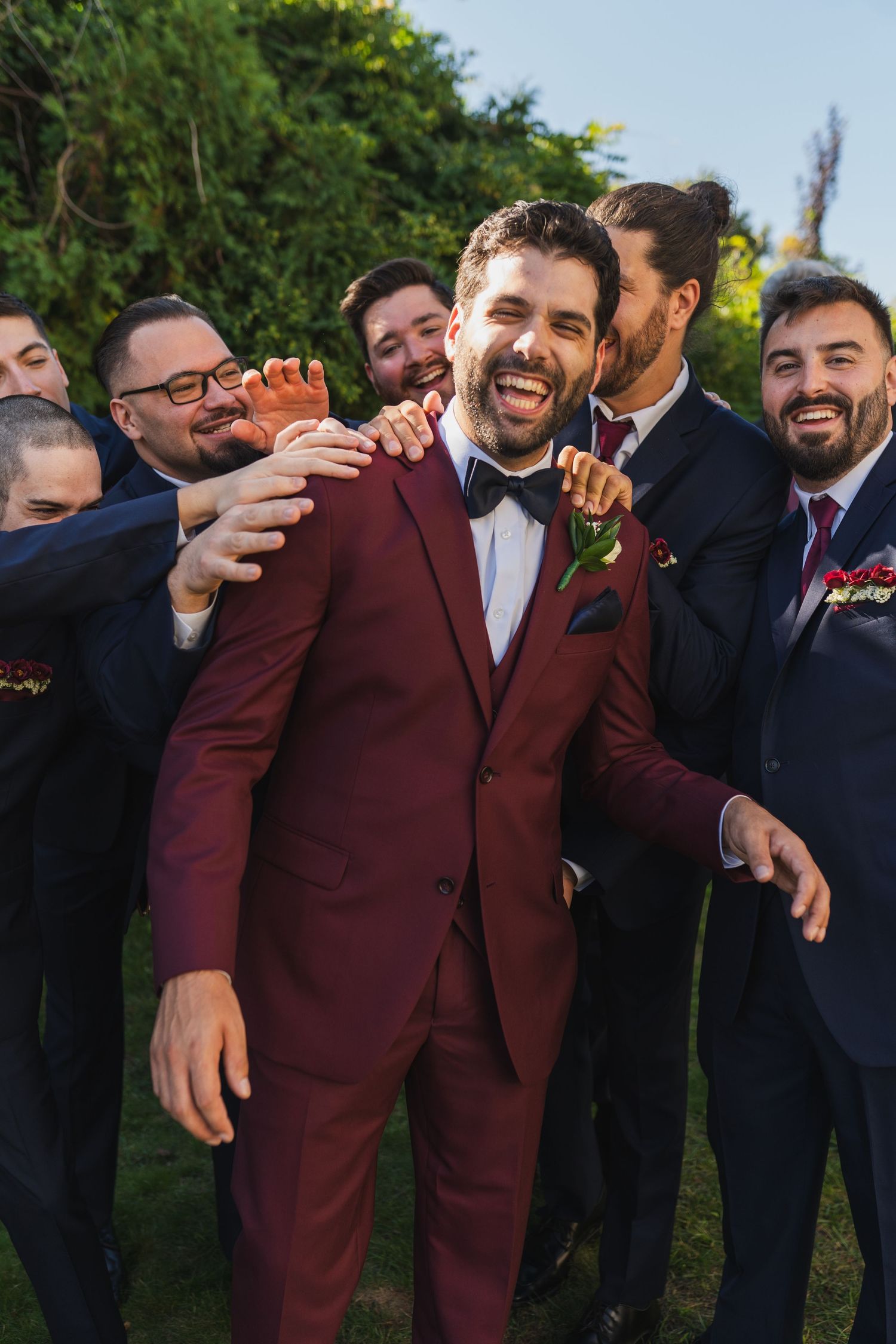 Wedding party members in burgundy and navy suits joke and laugh together during candid outdoor photo session.