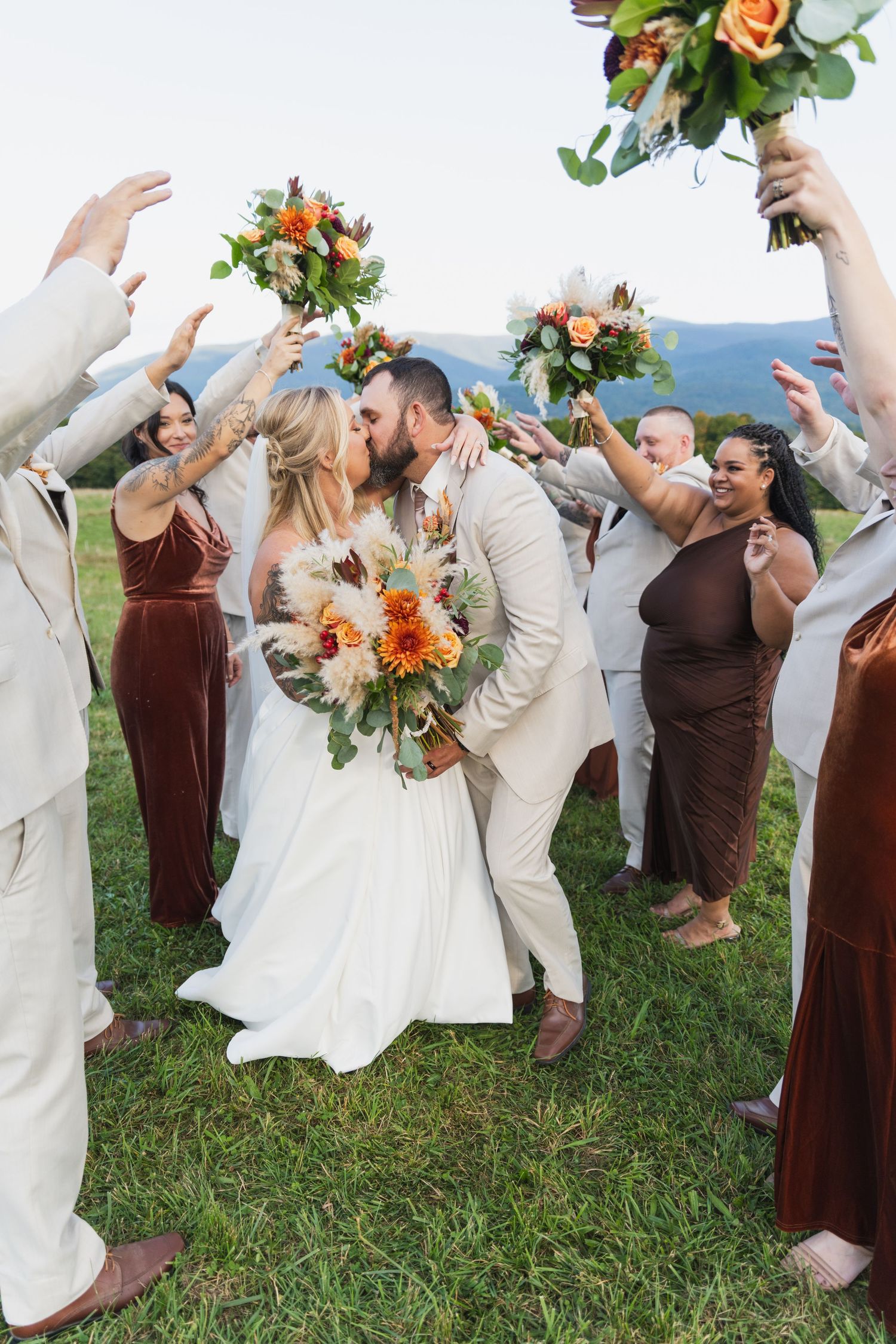 Wedding party in burgundy and cream outfits dancing joyfully on a grassy lawn with colorful floral bouquets.