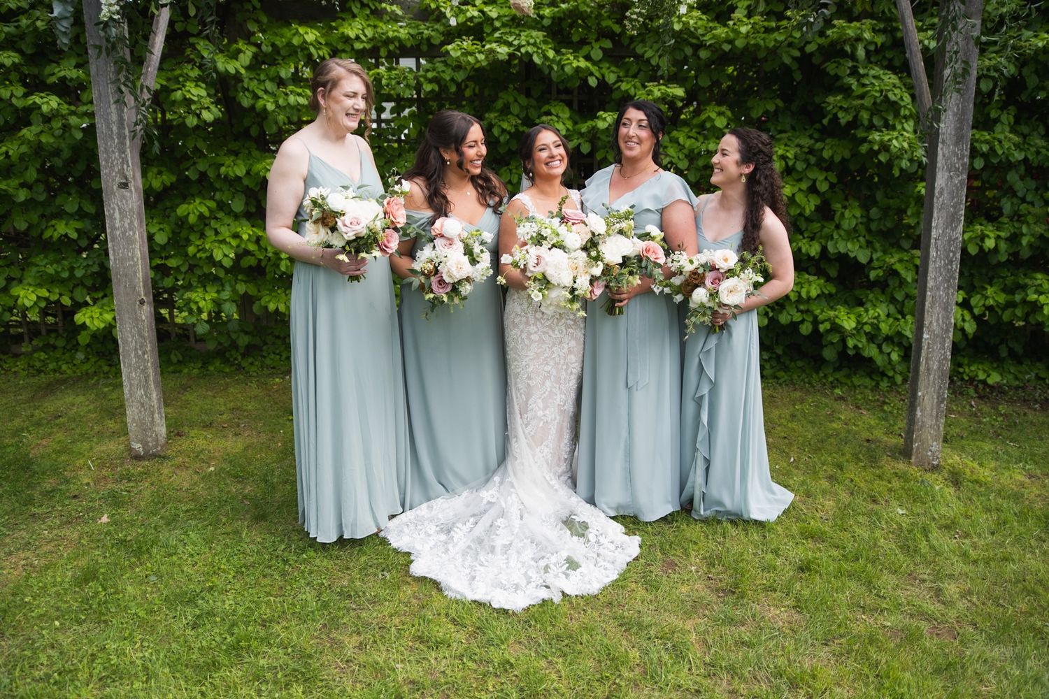 A bridal party in light blue dresses poses with bouquets in a garden setting.