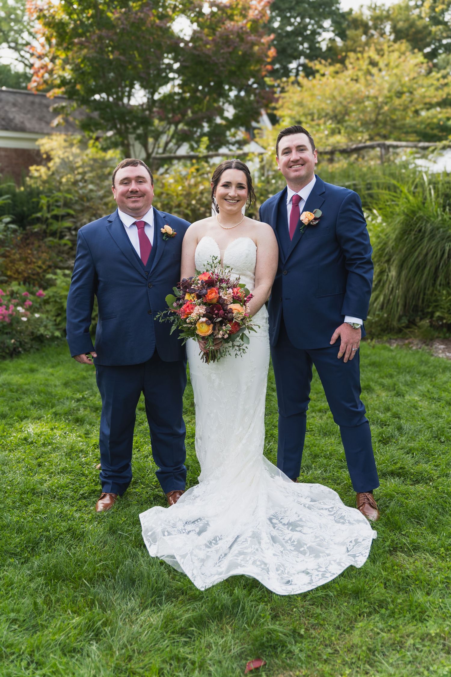 Wedding party in navy suits and white lace dress pose together on green lawn with autumn foliage in background.