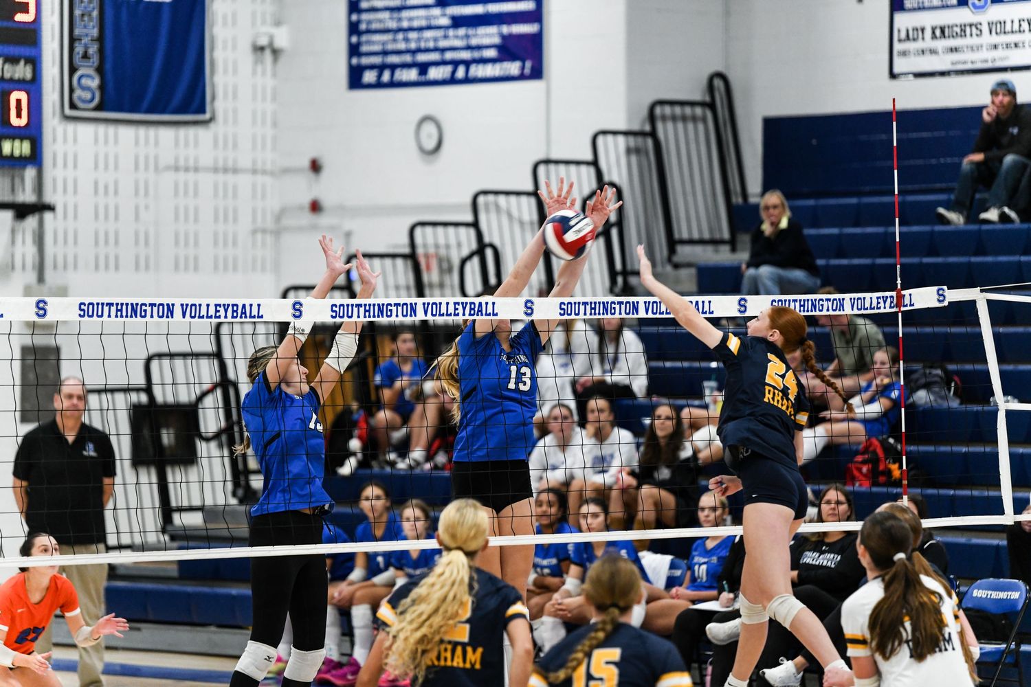 High school girls volleyball match with players in blue and yellow uniforms jumping at the net during an intense rally.