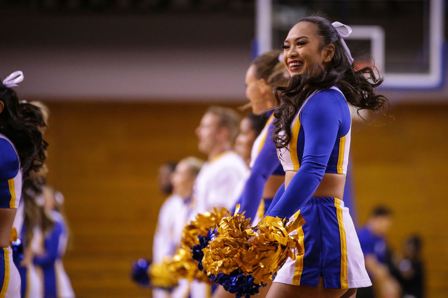 Cheerleader in blue and gold uniform smiling while holding gold pom-poms at an indoor basketball game.