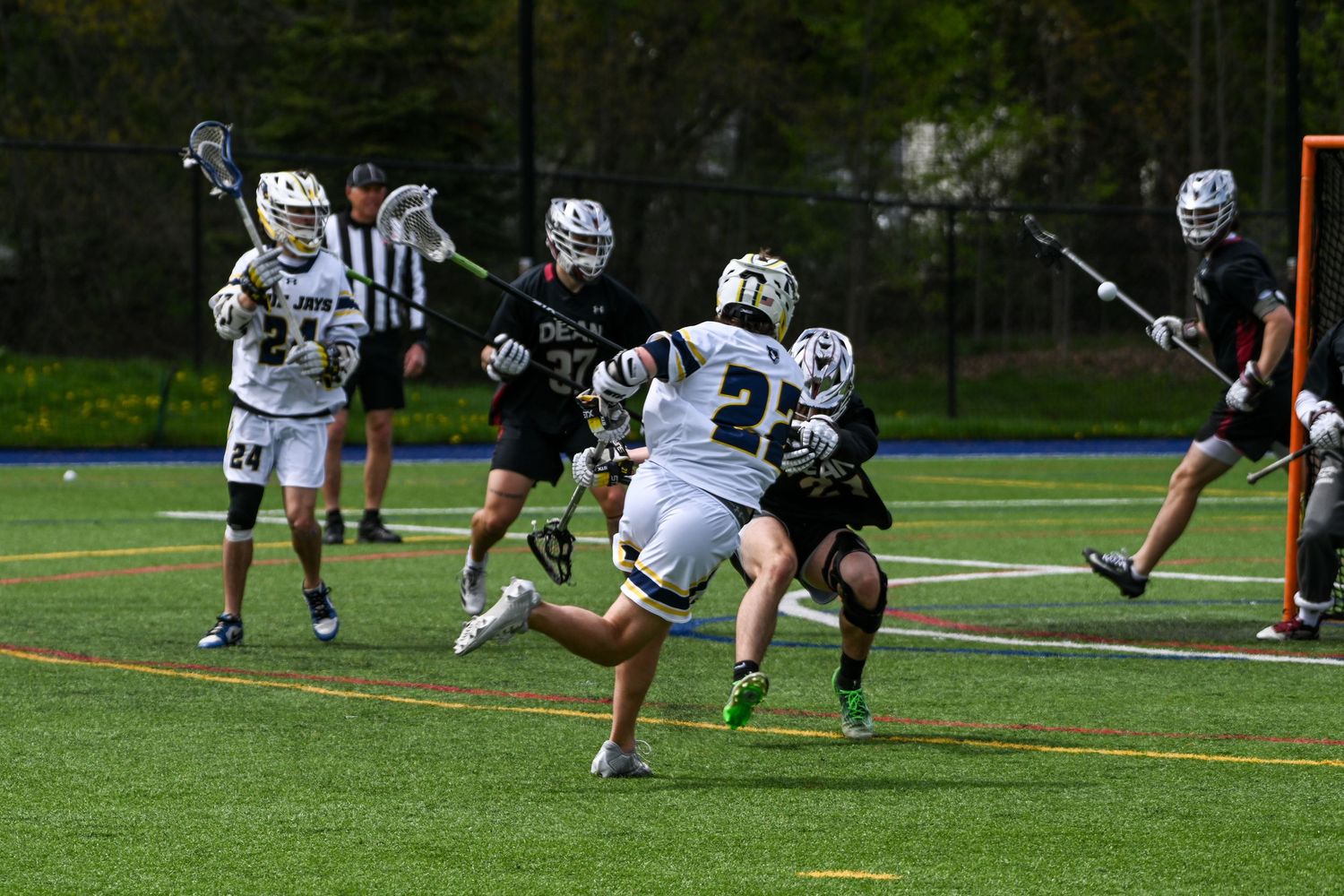 Youth lacrosse players compete on a turf field, with one player driving toward the goal while defenders attempt to block.