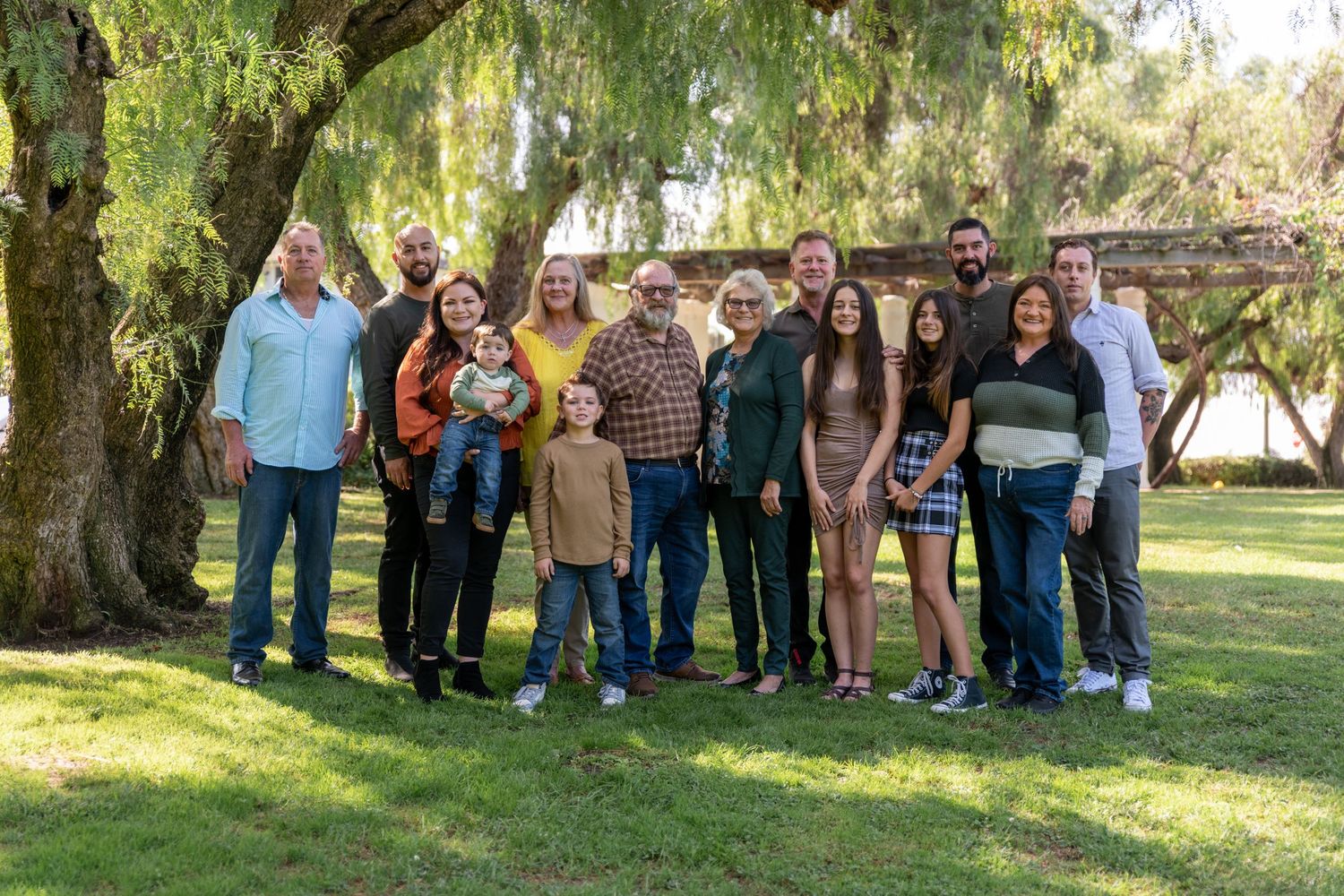 Large extended family gathering posed under weeping willow trees in a park setting.
