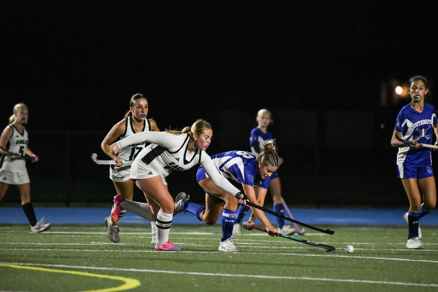 Female field hockey players compete for the ball during a night game, with players in white and blue uniforms.