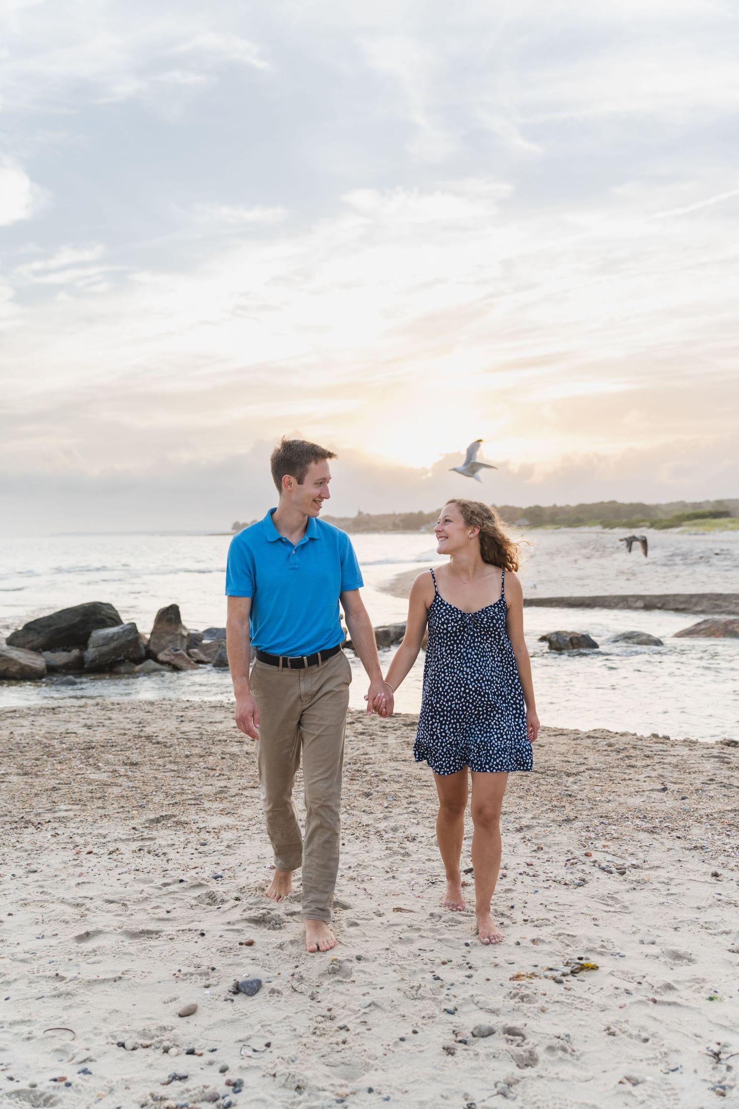 Couple taking a romantic stroll along a sandy beach with rocks and seagulls during sunset.