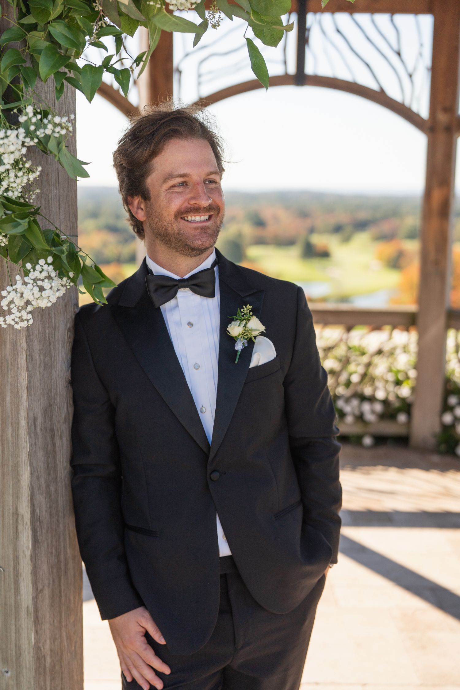 A groom in a classic black tuxedo and bow tie smiles while standing in a rustic wooden pavilion.