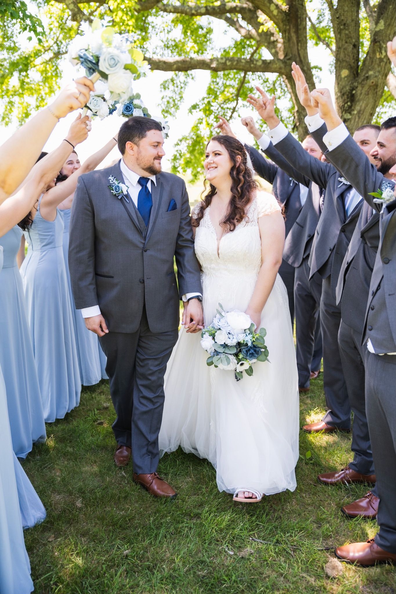 Wedding party in light blue dresses and dark suits celebrating with raised arms under a tree.