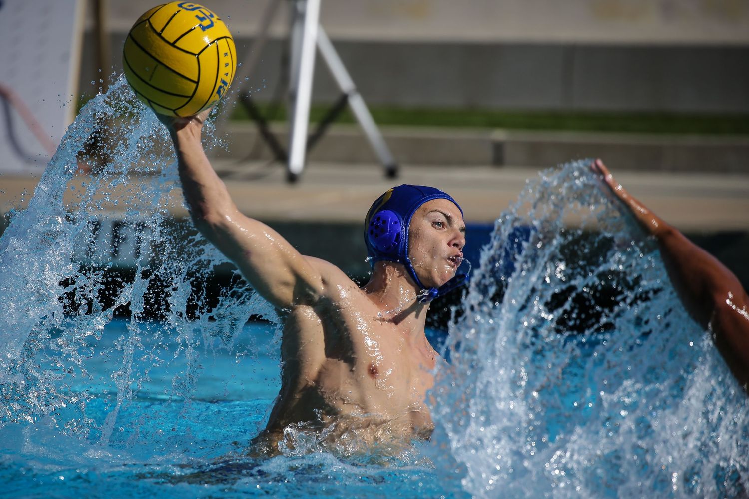 Water polo player rises from pool to throw yellow ball during competitive match outdoors.