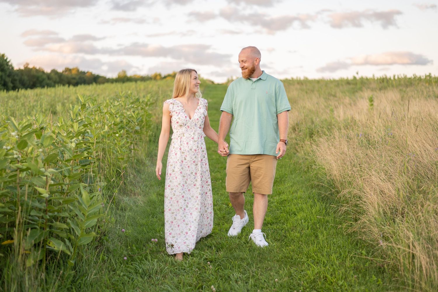 Couple walks hand in hand through tall grass field during golden hour.