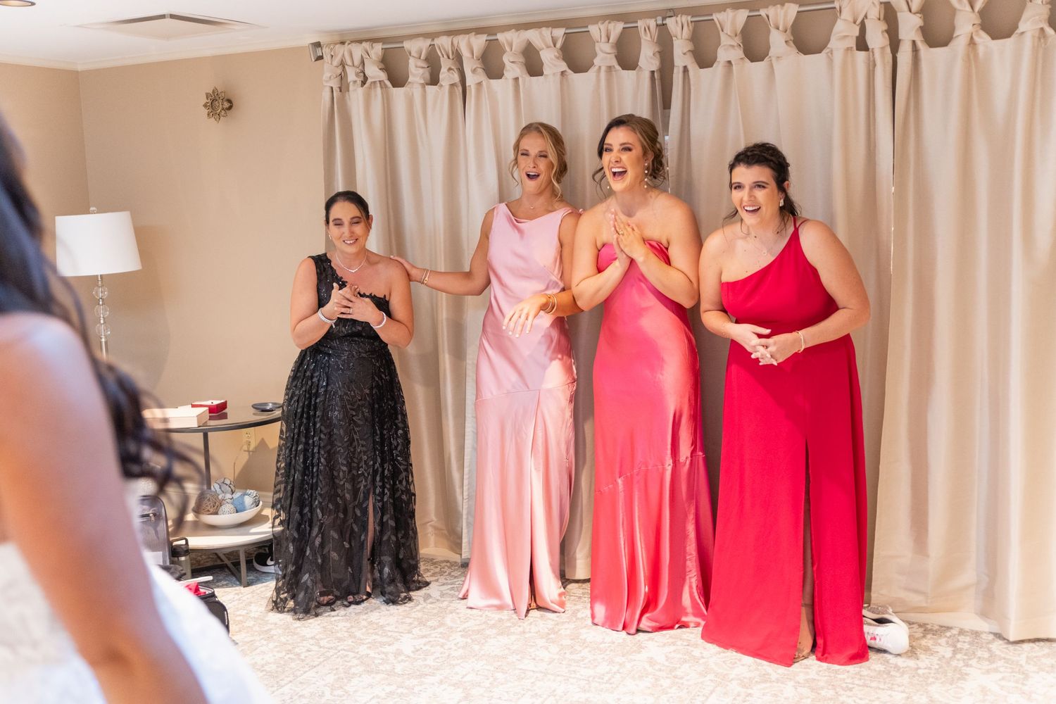 A group in formal evening gowns reacts with joy and excitement during a special occasion in a room with beige curtains.