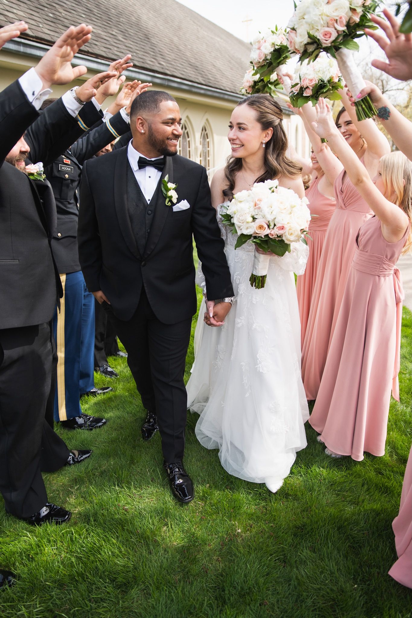 Wedding couple walking under raised arms of guests in pink dresses.