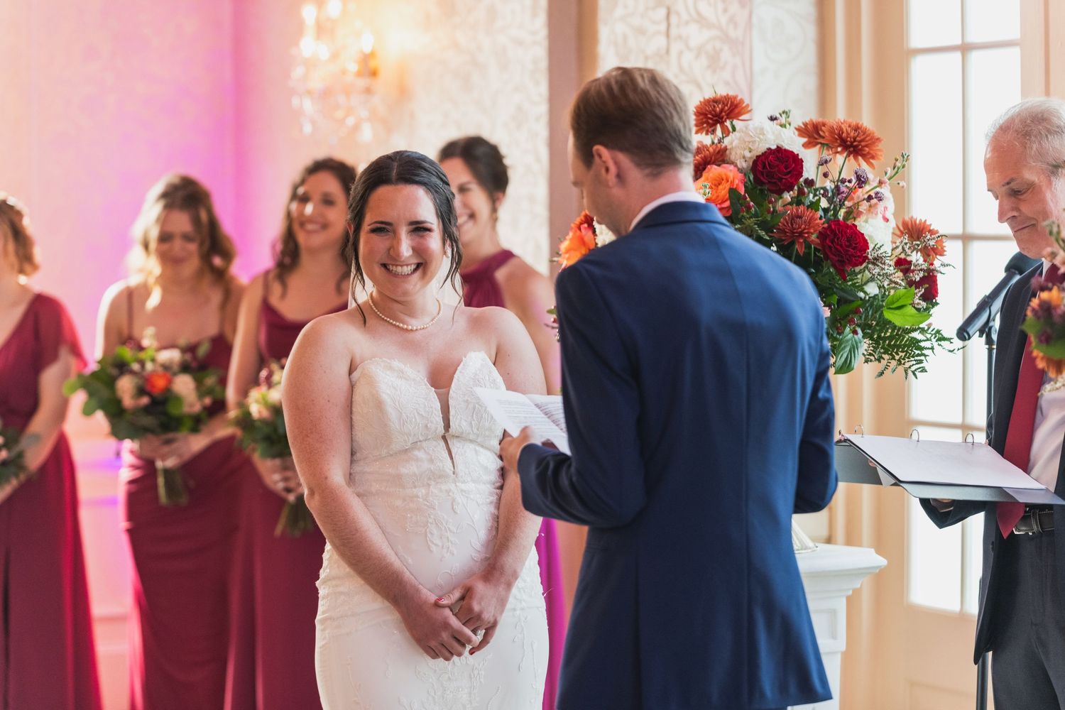 Wedding guests celebrate during a reception in an elegant ballroom with pink lighting and large windows.