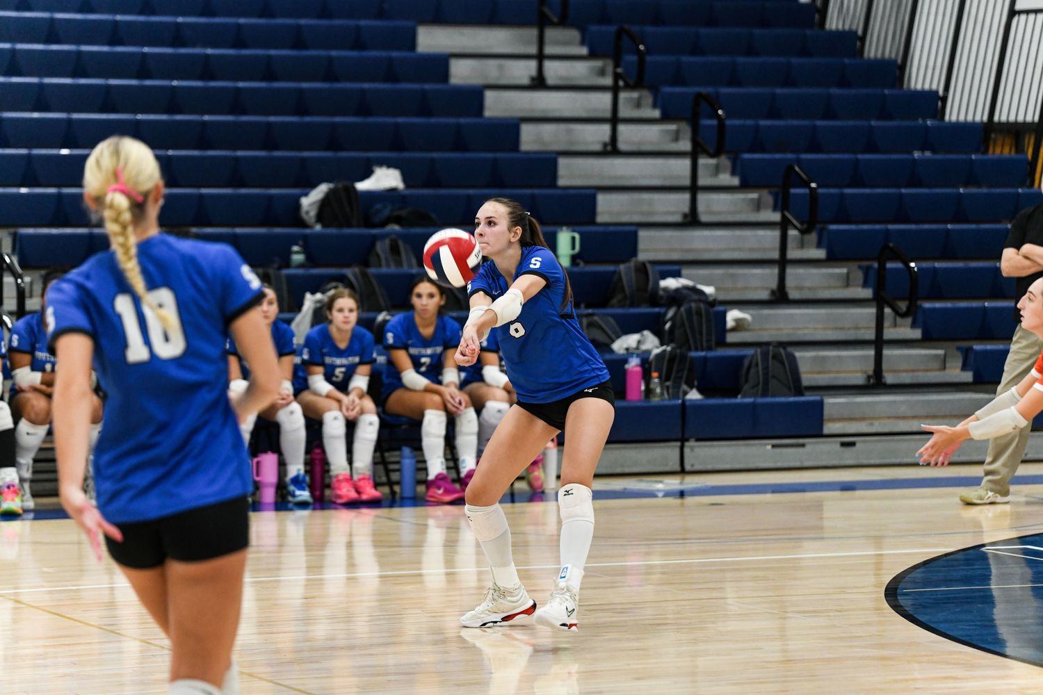 Female volleyball player in blue uniform performs a dig during an indoor match with teammates on the court.