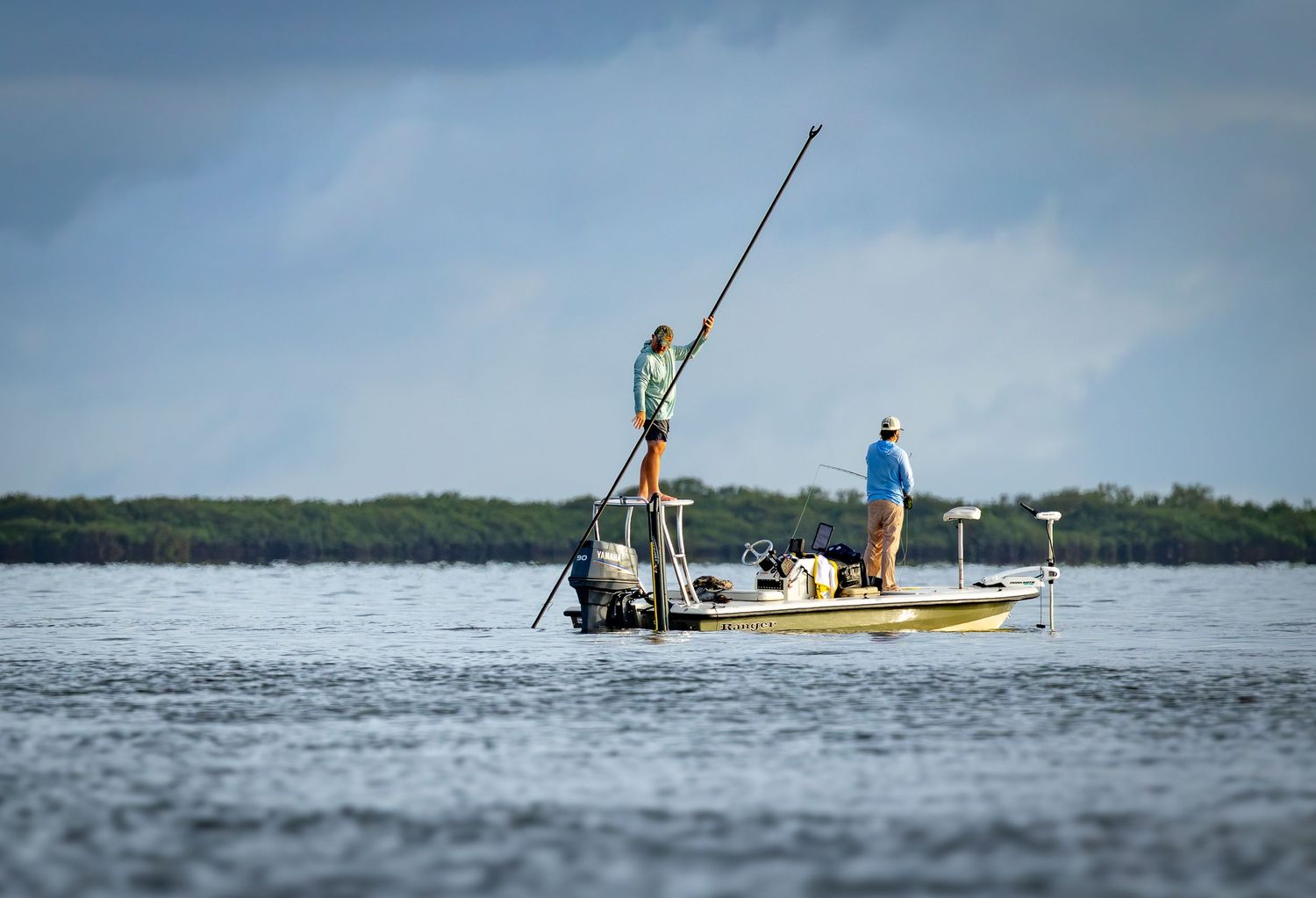 A guide poles a shallow-water skiff while an angler watches the flats.