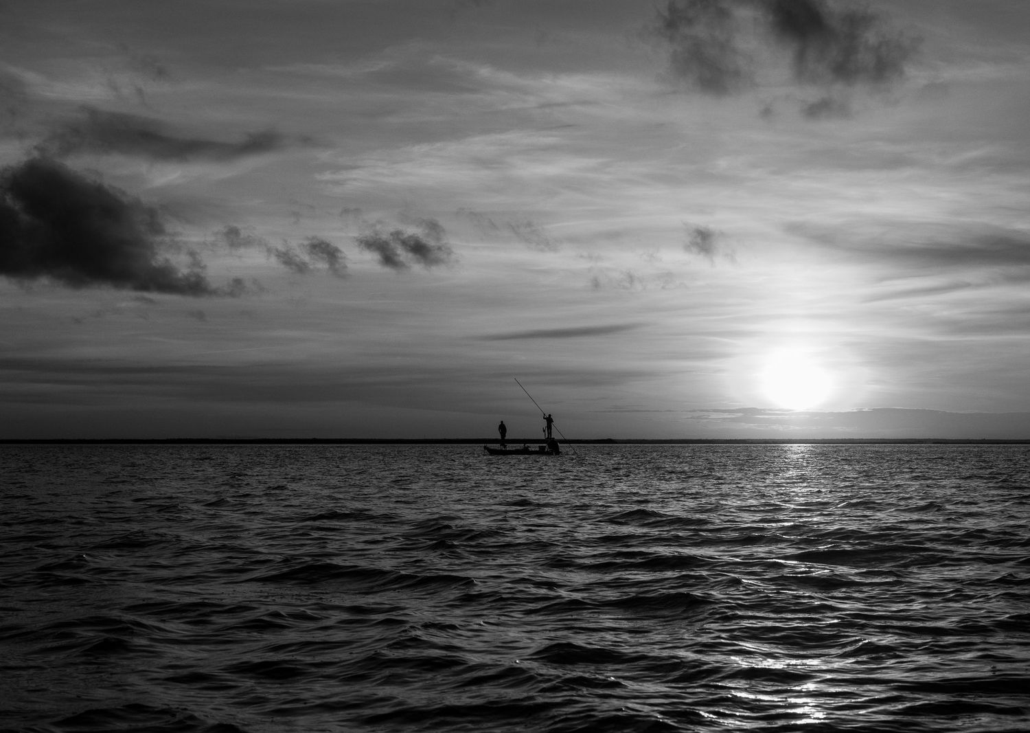 A distant flats skiff crosses rippling water beneath a luminous black-and-white sunrise.