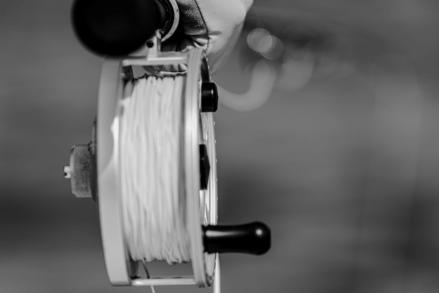 A fly reel, pale backing, and handle fill a close black-and-white composition.