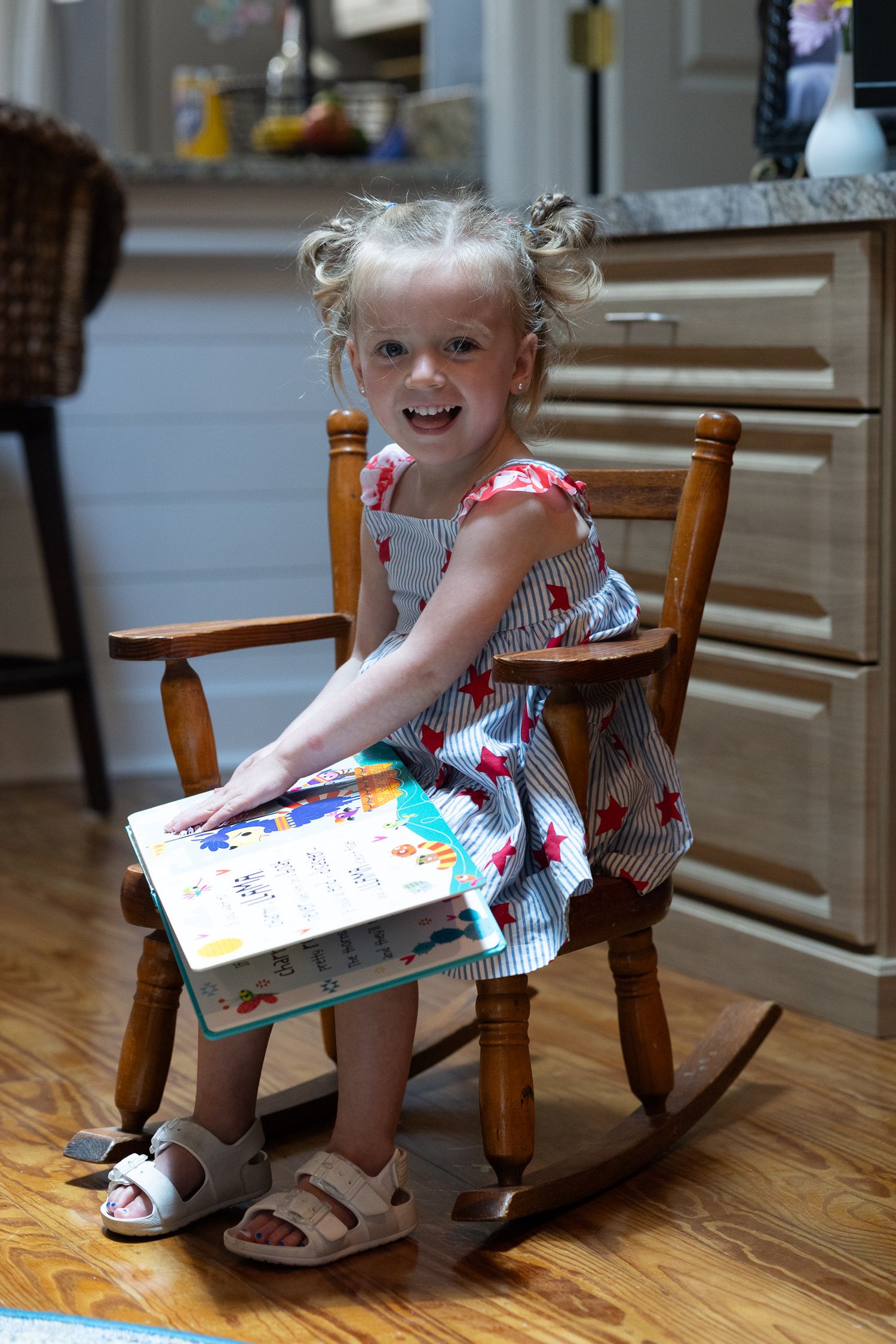 A young child smiles from a small wooden rocking chair with an open picture book.