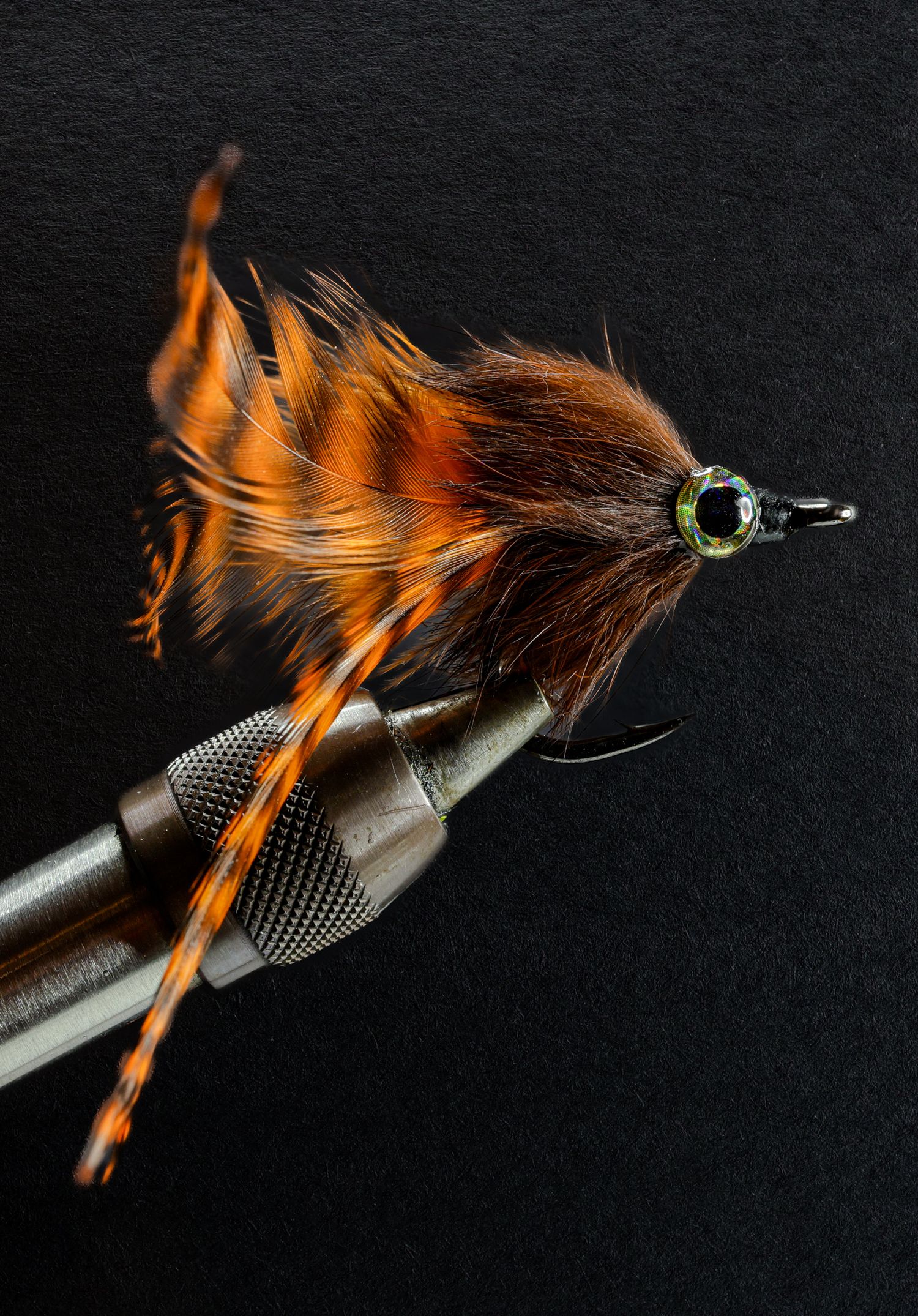 Orange fly
Macro photograph of an orange hand-tied saltwater fly in a fly-tying vise, featuring bright feather movement, detailed tying mate